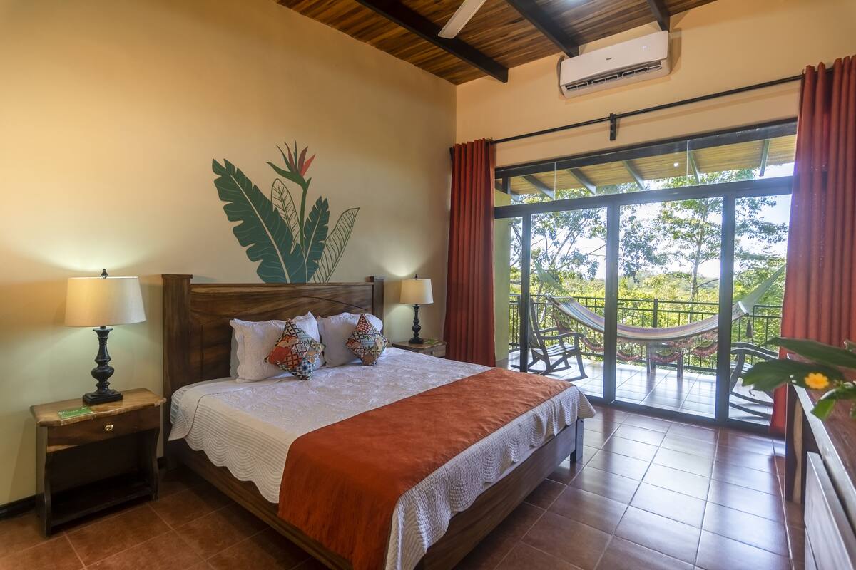 Une chambre à coucher avec un lit, des tables de chevet, une grande fenêtre et une vue sur l’extérieur.