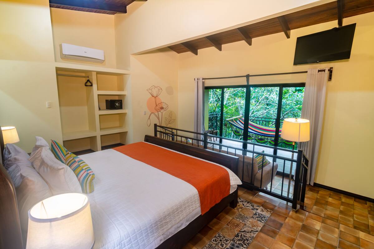 Une chambre avec un lit, des lampes de chevet, une armoire, une télévision et un balcon donnant sur un espace verdoyant.