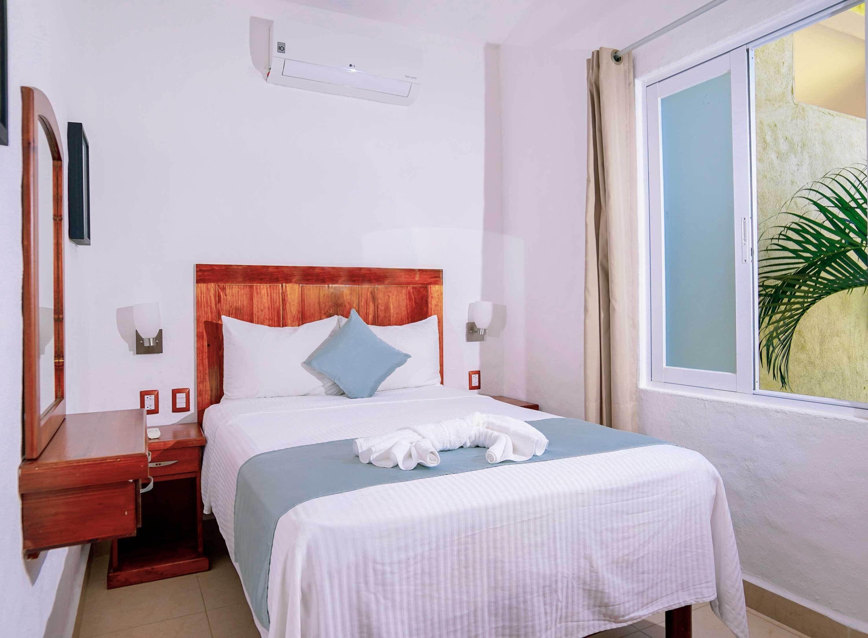 Habitación de hotel con cabecera de madera, cama con ropa de cama blanca y azul, mesita de noche, espejo, aire acondicionado instalado en la pared y ventana con vistas a zonas verdes.