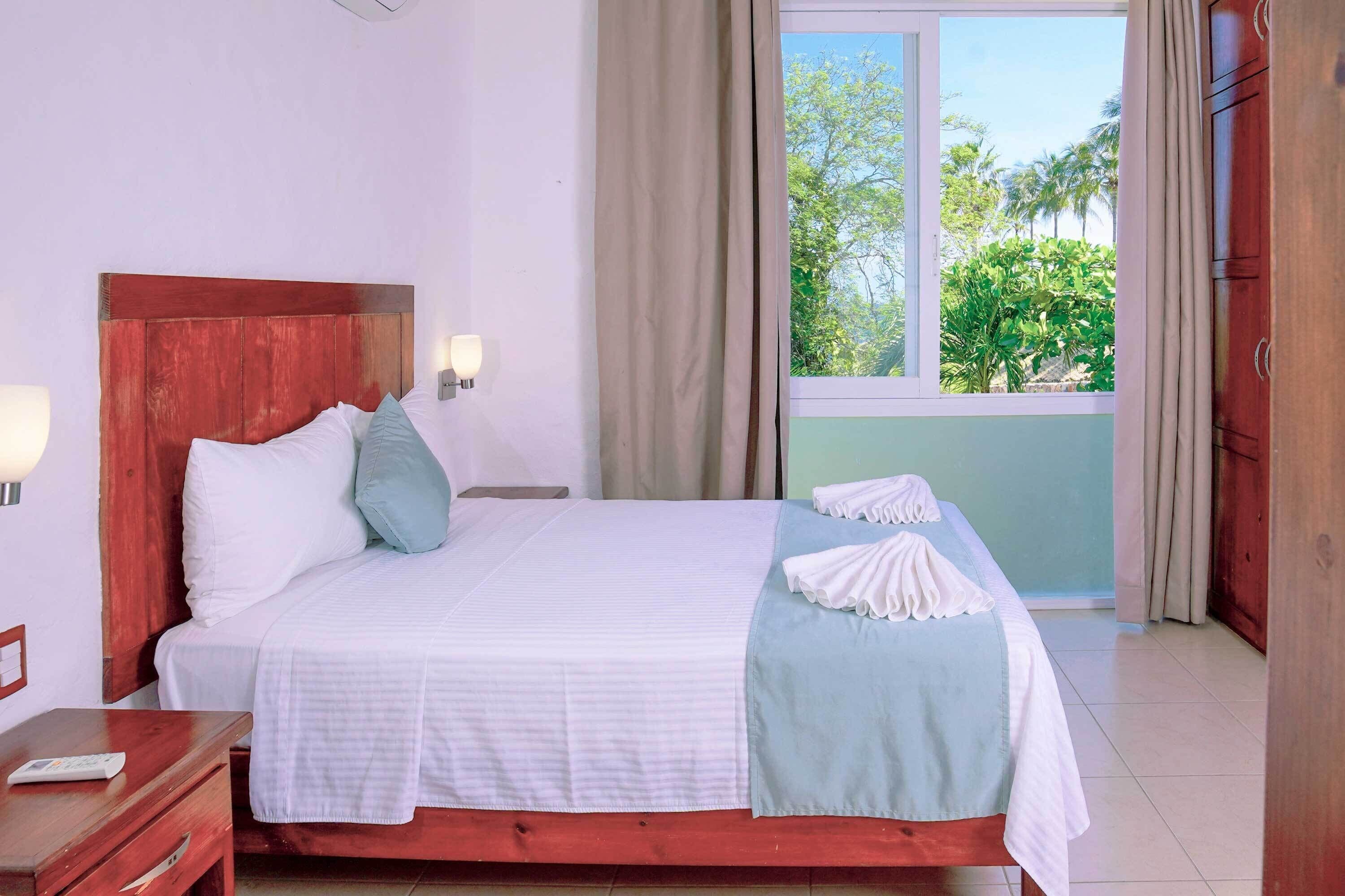 A neatly made bed with white and light blue bedding, a wooden headboard, and a view of greenery outside.
