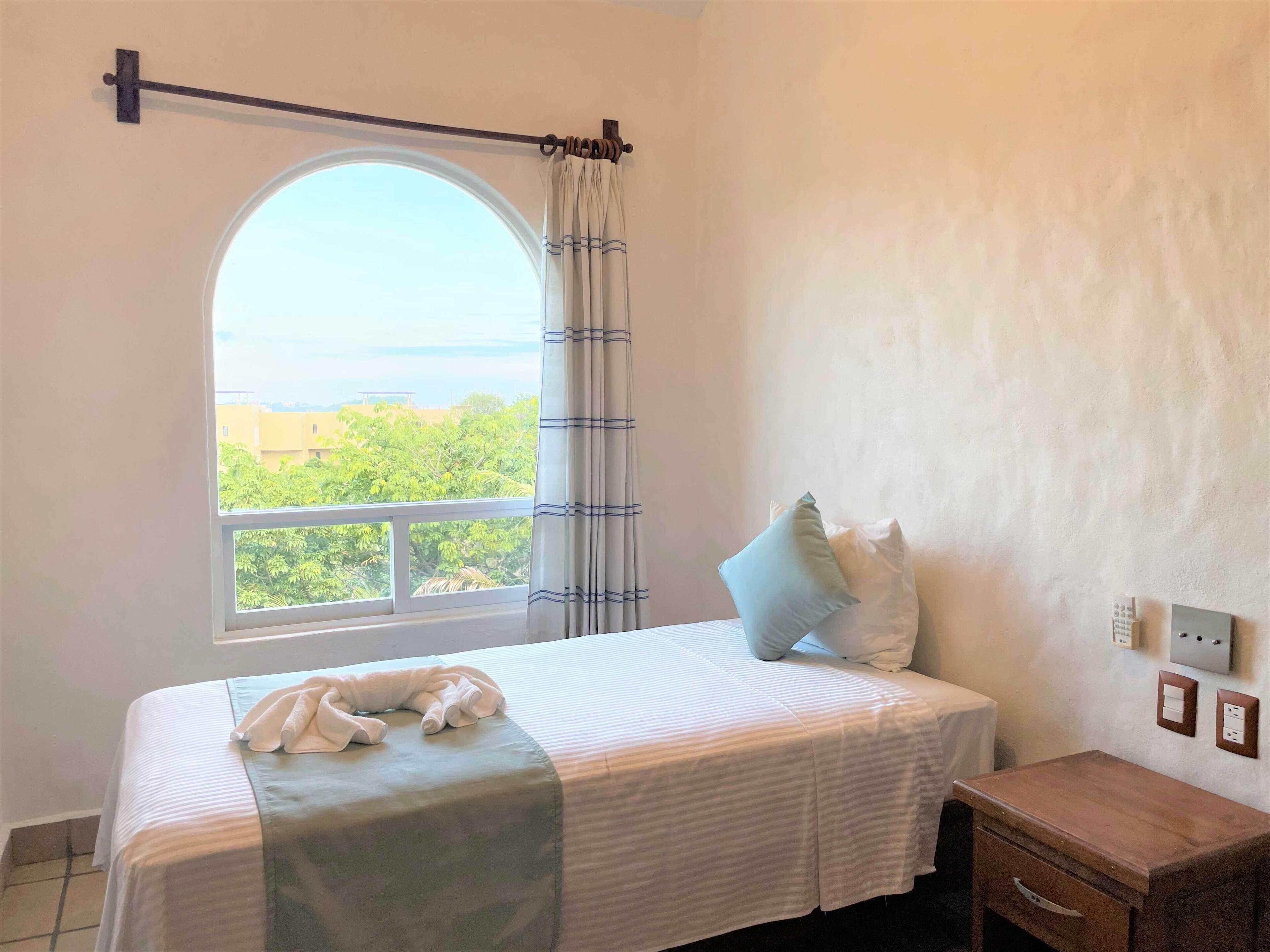 A bedroom with a bed, a nightstand, a wooden dresser, a window with curtains, and a view of greenery outside.