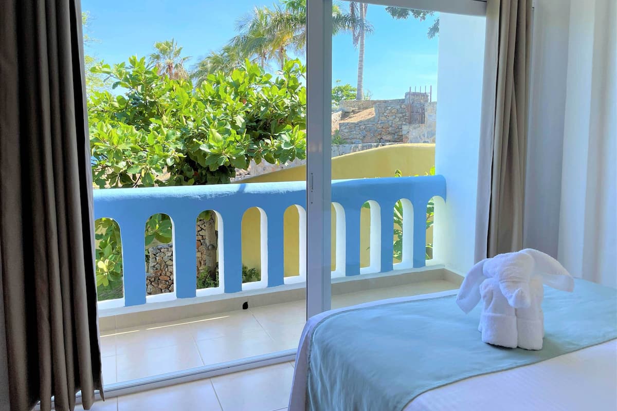Une chambre d’hôtel avec une grande fenêtre, un lit recouvert d’une couverture blanche et une vue sur un balcon bleu orné de plantes.