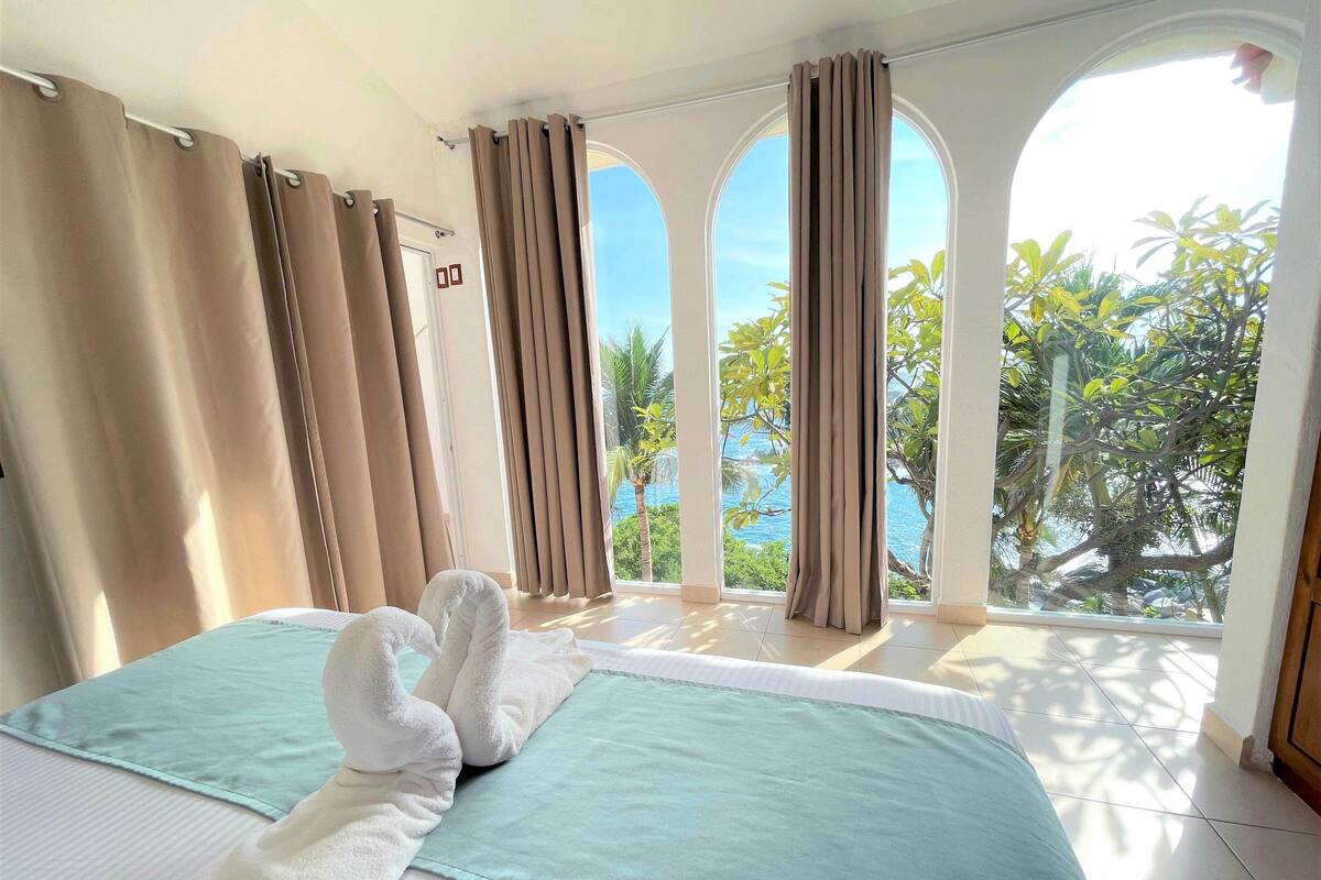 Une chambre d’hôtel avec deux lits, des rideaux de couleur beige et une vue sur les palmiers et la mer.