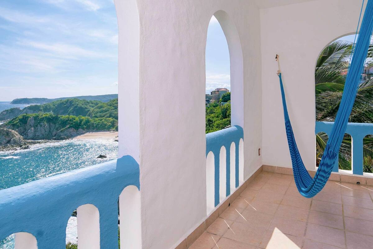 Un balcon avec vue sur l’océan et les falaises, un hamac bleu et une balustrade blanche.