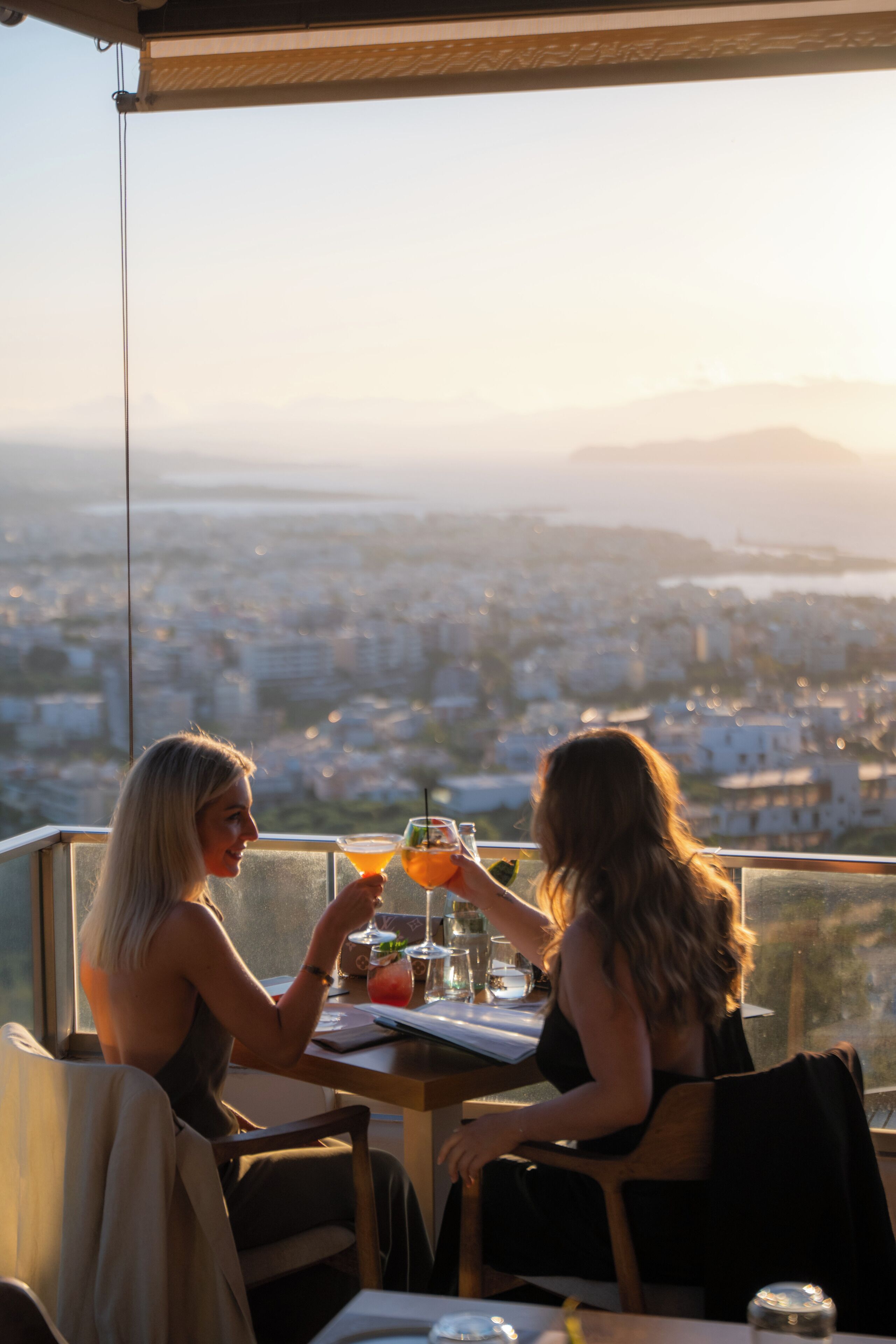 Deux personnes sont assises à une table, devant des boissons, et elles ont une vue sur un paysage urbain avec des montagnes à l'horizon.