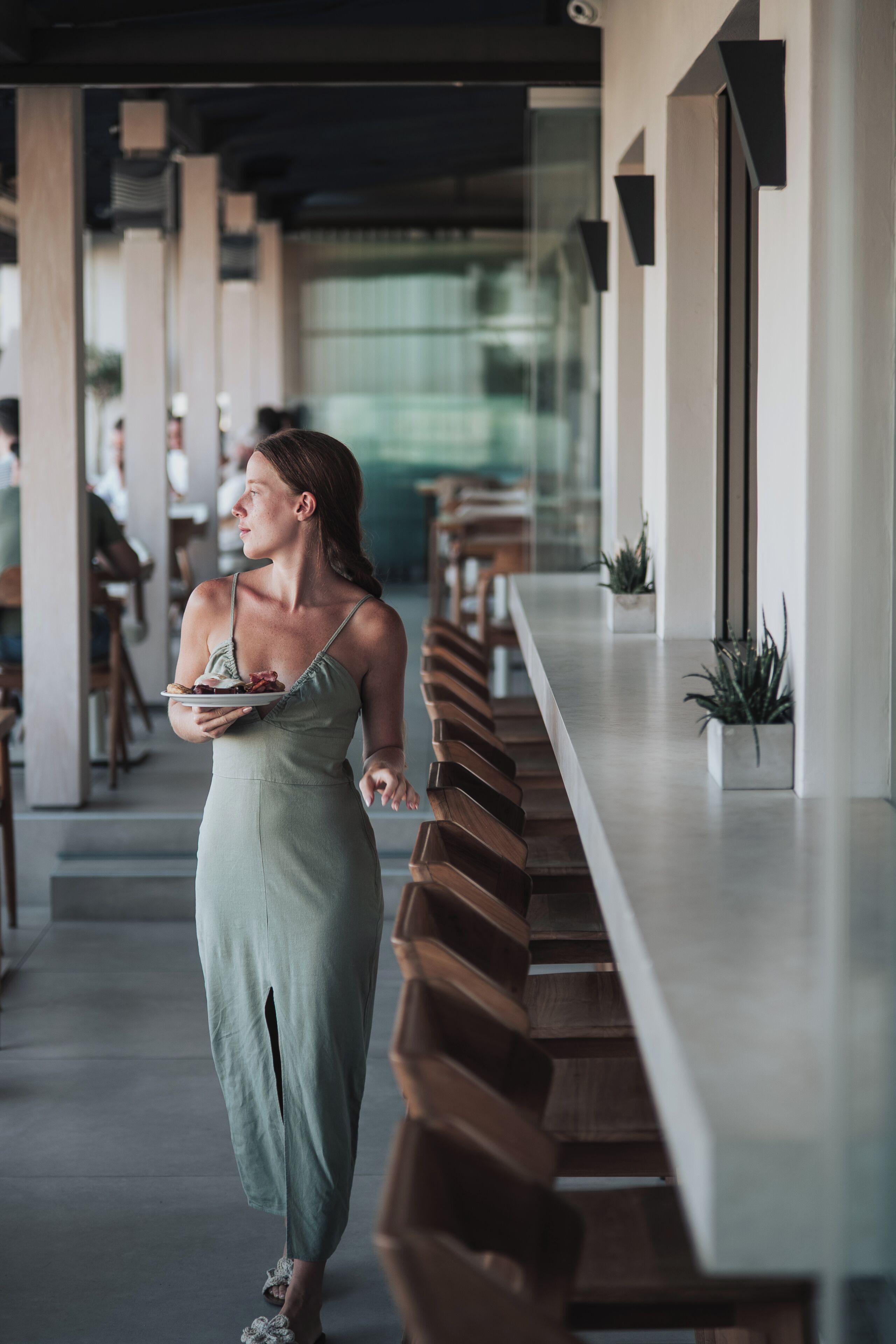 A woman walking in a modern interior with a long bench, a glass partition, and potted plants.