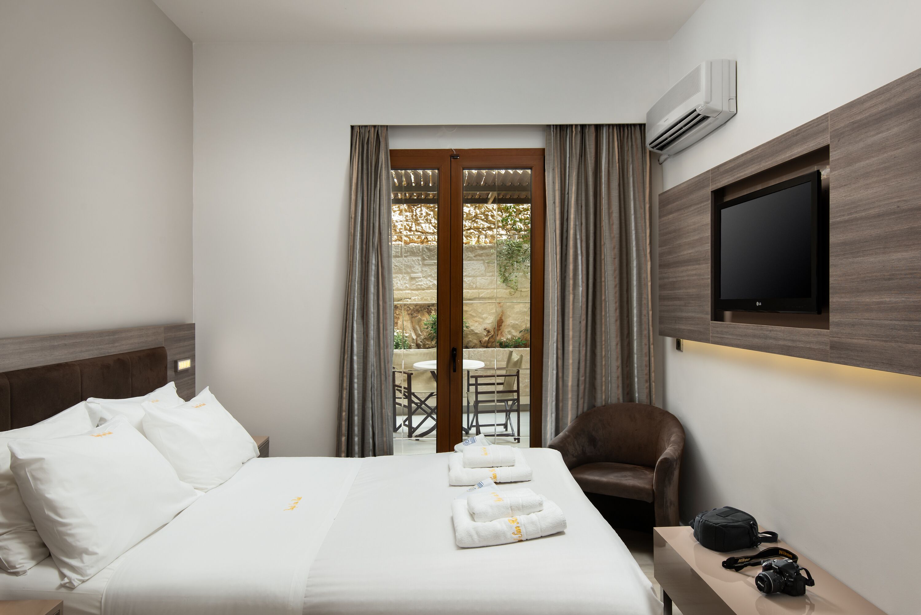 A hotel room with a bed, a TV, a chair, and a table with a camera and a bag.