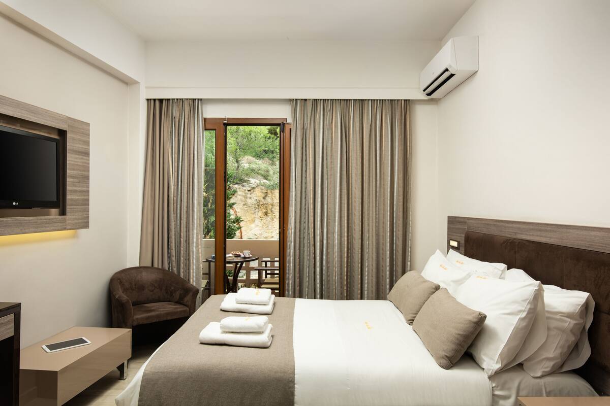 A hotel room with a bed, a TV, a desk, a chair, and a view of greenery outside.