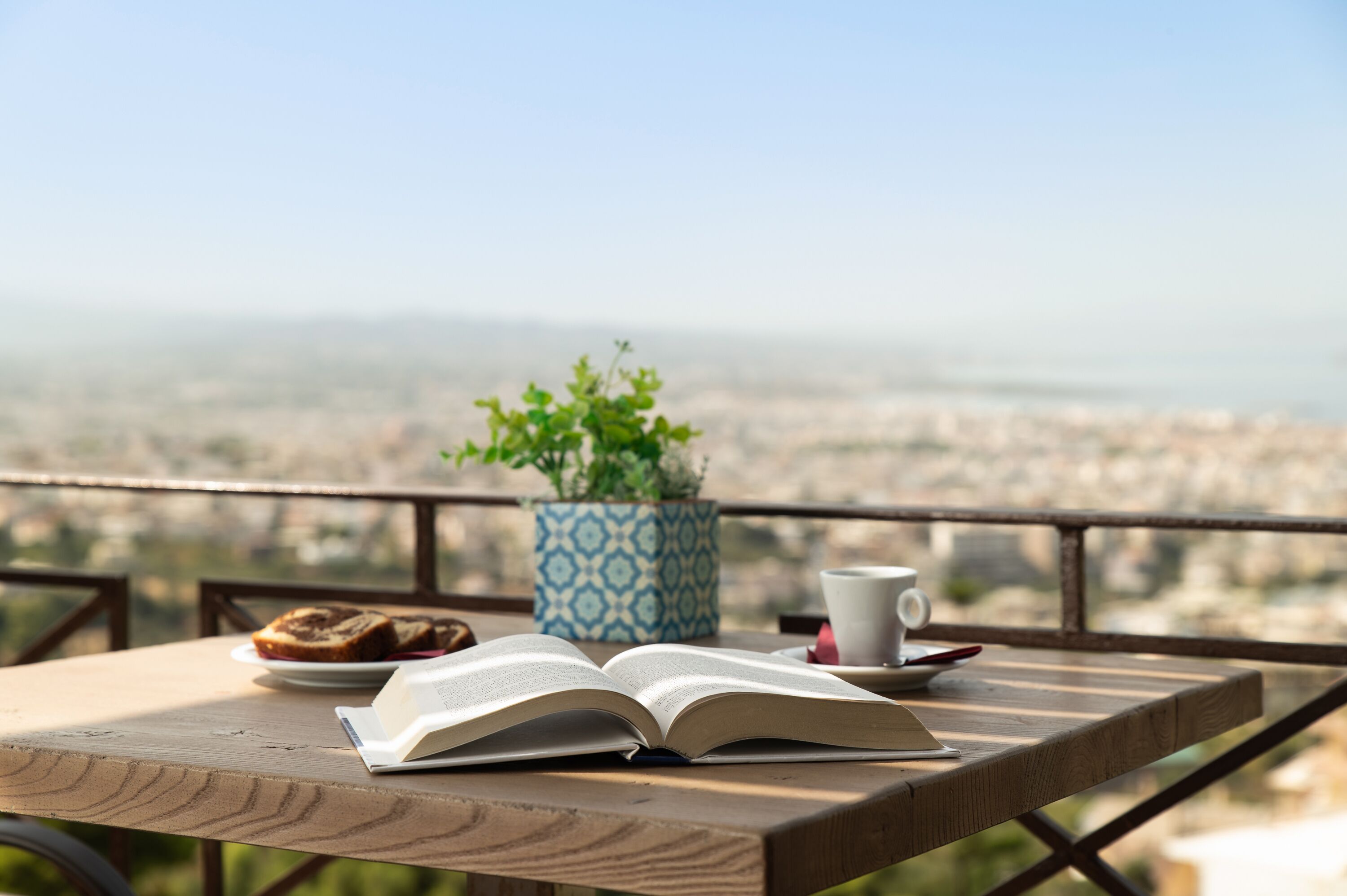 Ein Holztisch mit einem offenen Buch, einer Tasse und einem Teller mit Essen, vor einer schönen Stadtlandschaft.