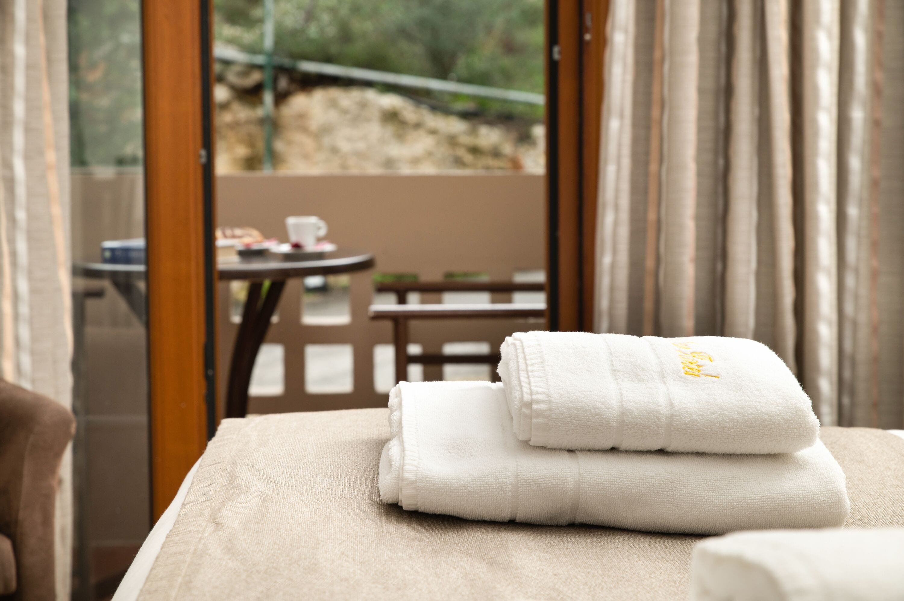 Una cama bien hecha con toallas blancas dobladas y vistas a una terraza exterior con una mesa y sillas.