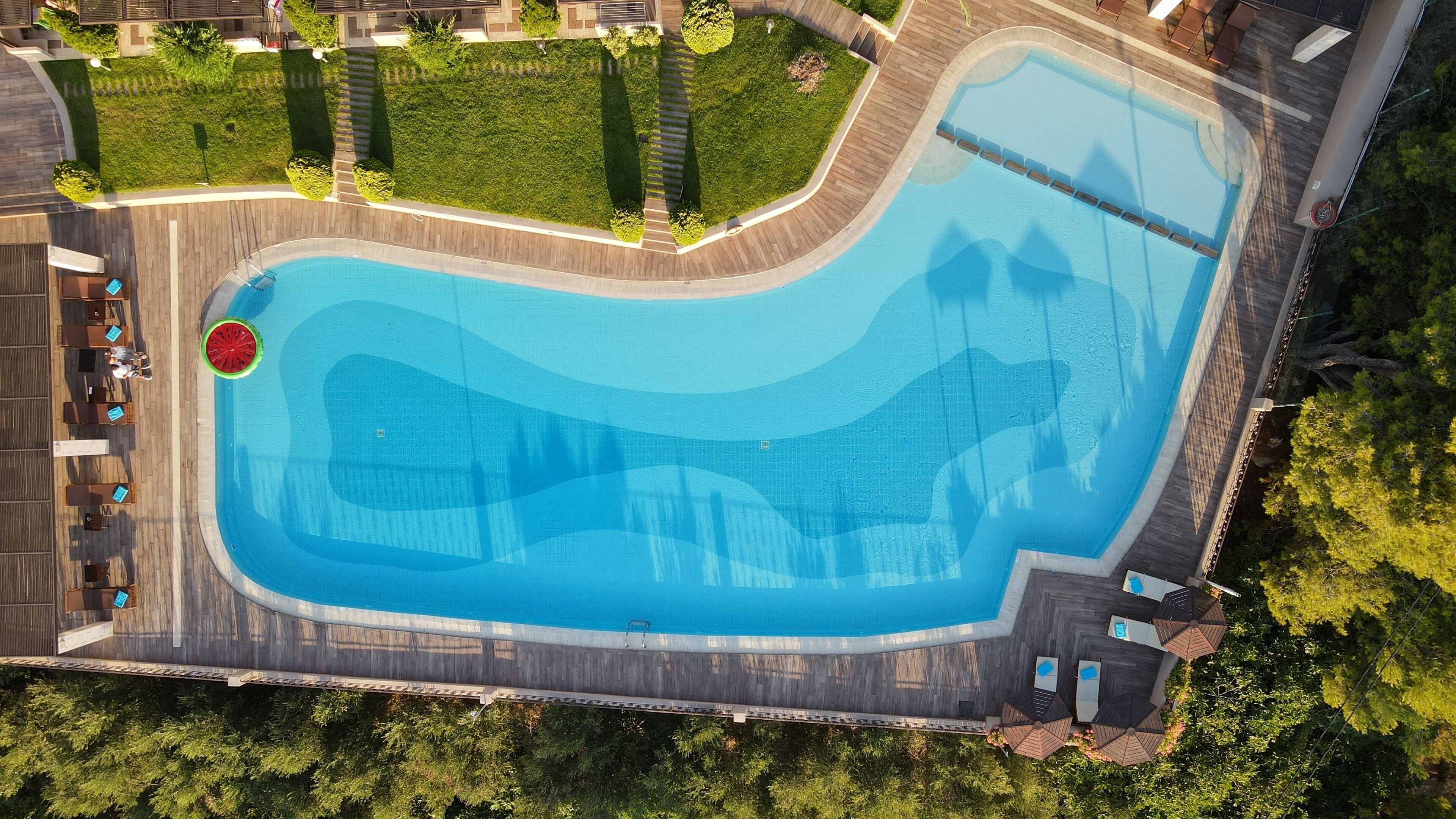 An aerial view of a swimming pool with a wooden deck surrounding it, equipped with lounge chairs and umbrellas.