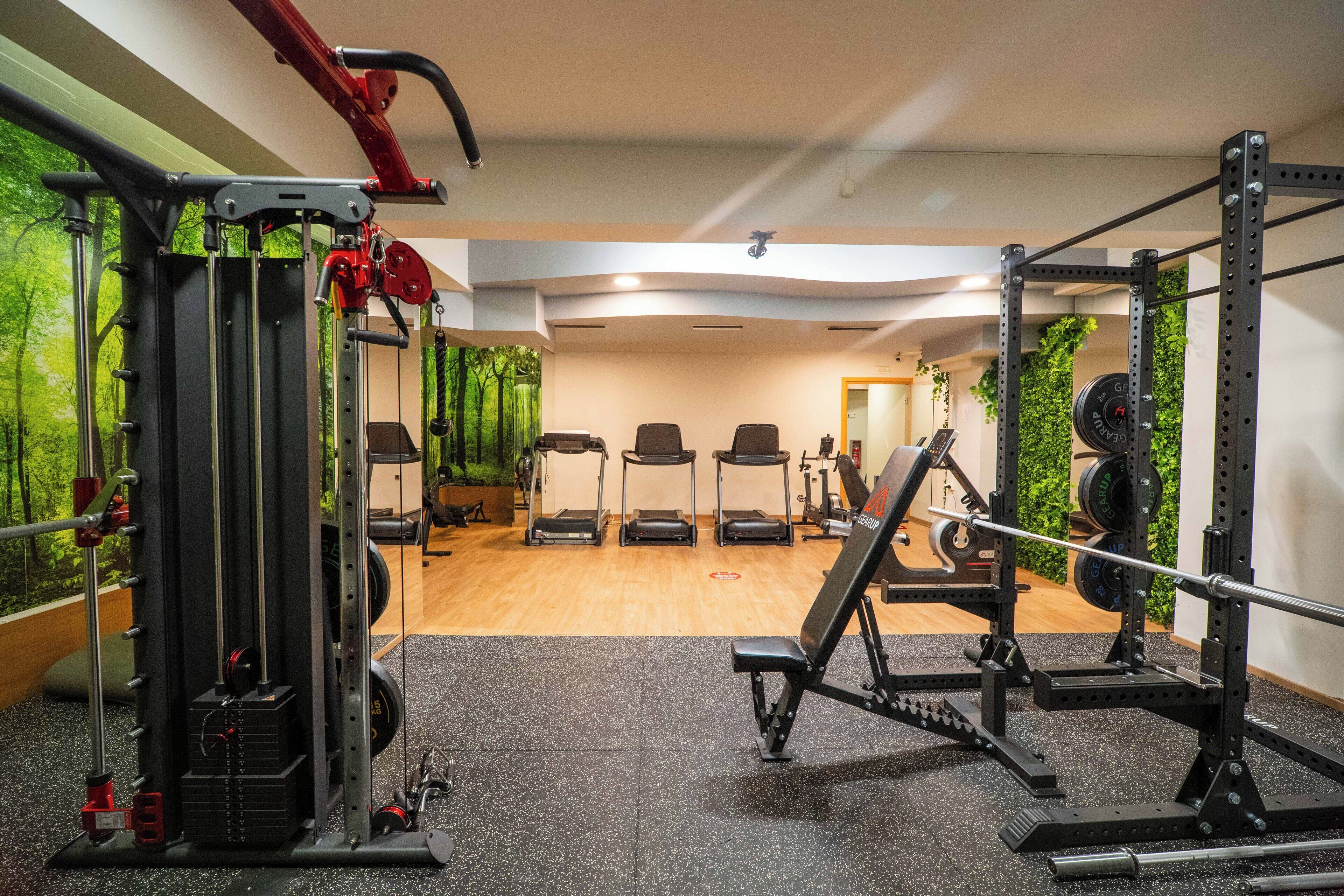 A gym with weightlifting equipment, treadmills, and a view of greenery through large windows.