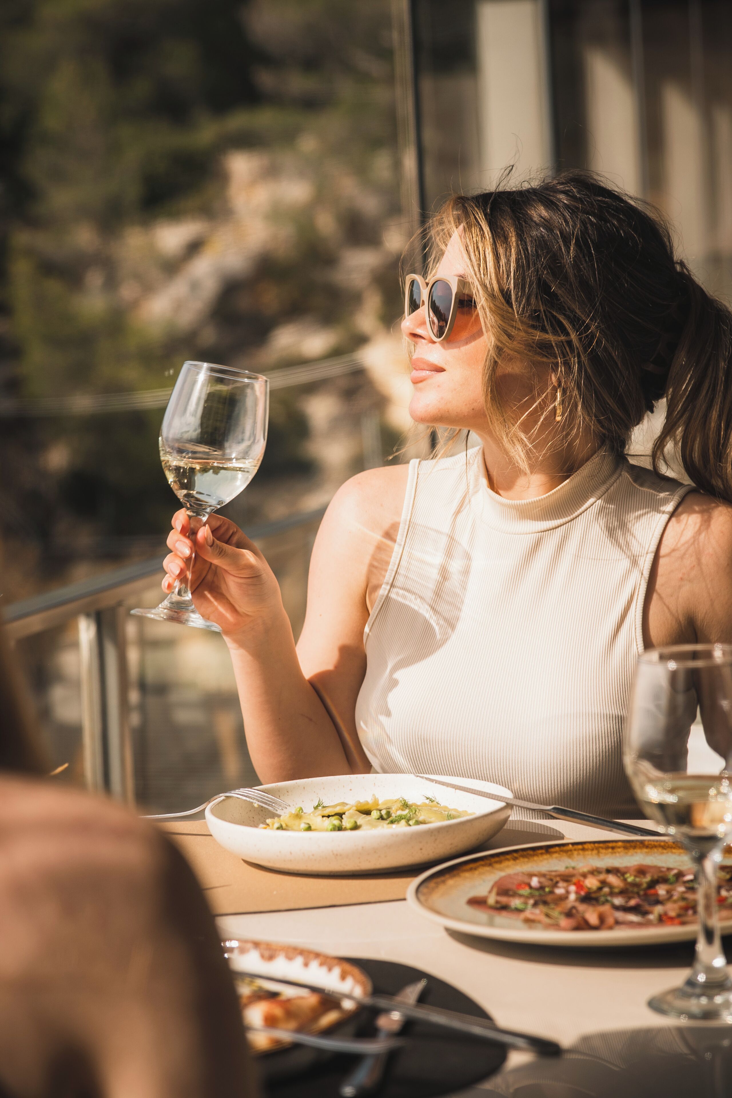 A person seated at a table with a glass of white wine, plates of food, and a window with a view of greenery.