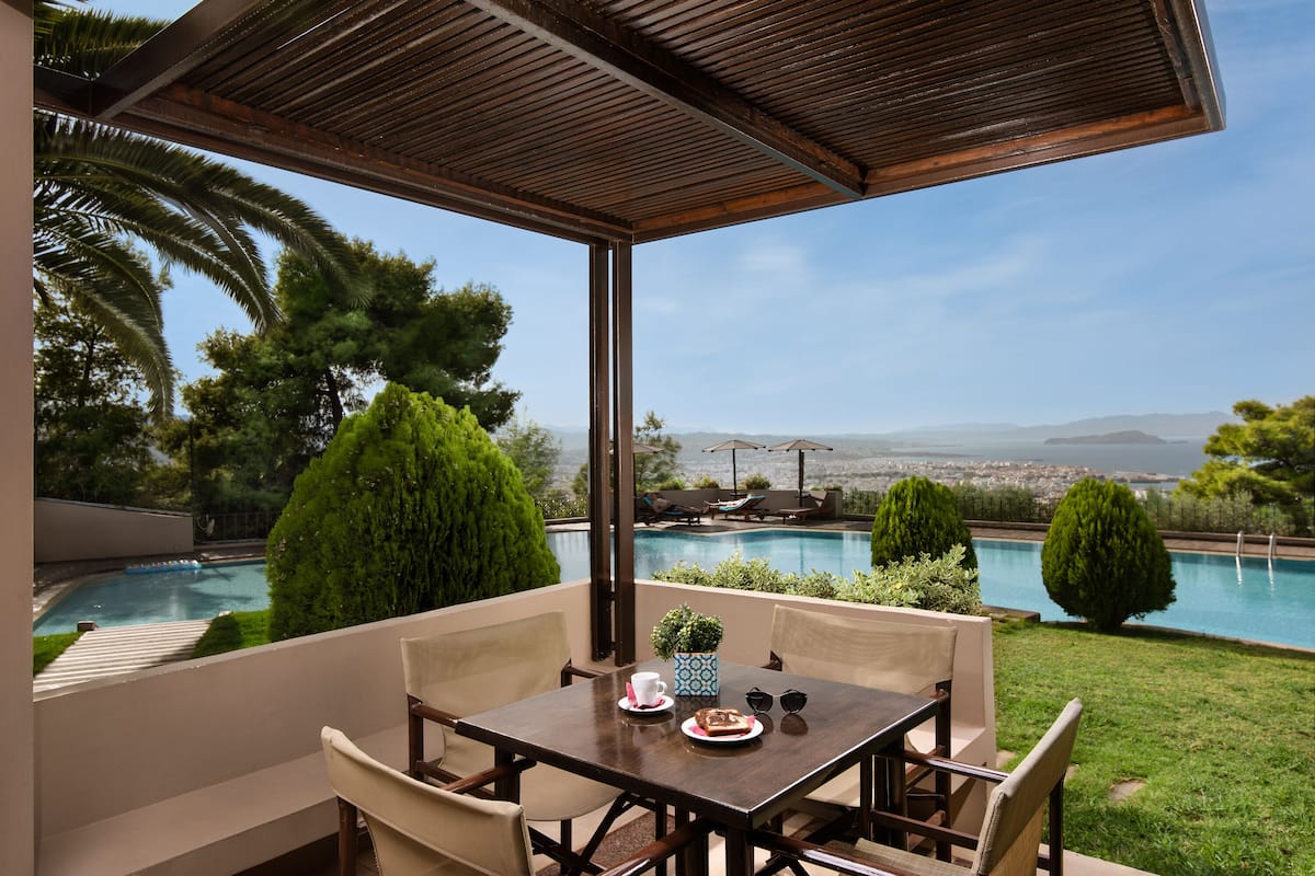 A covered outdoor seating area with a table and chairs, overlooking a swimming pool and a cityscape.