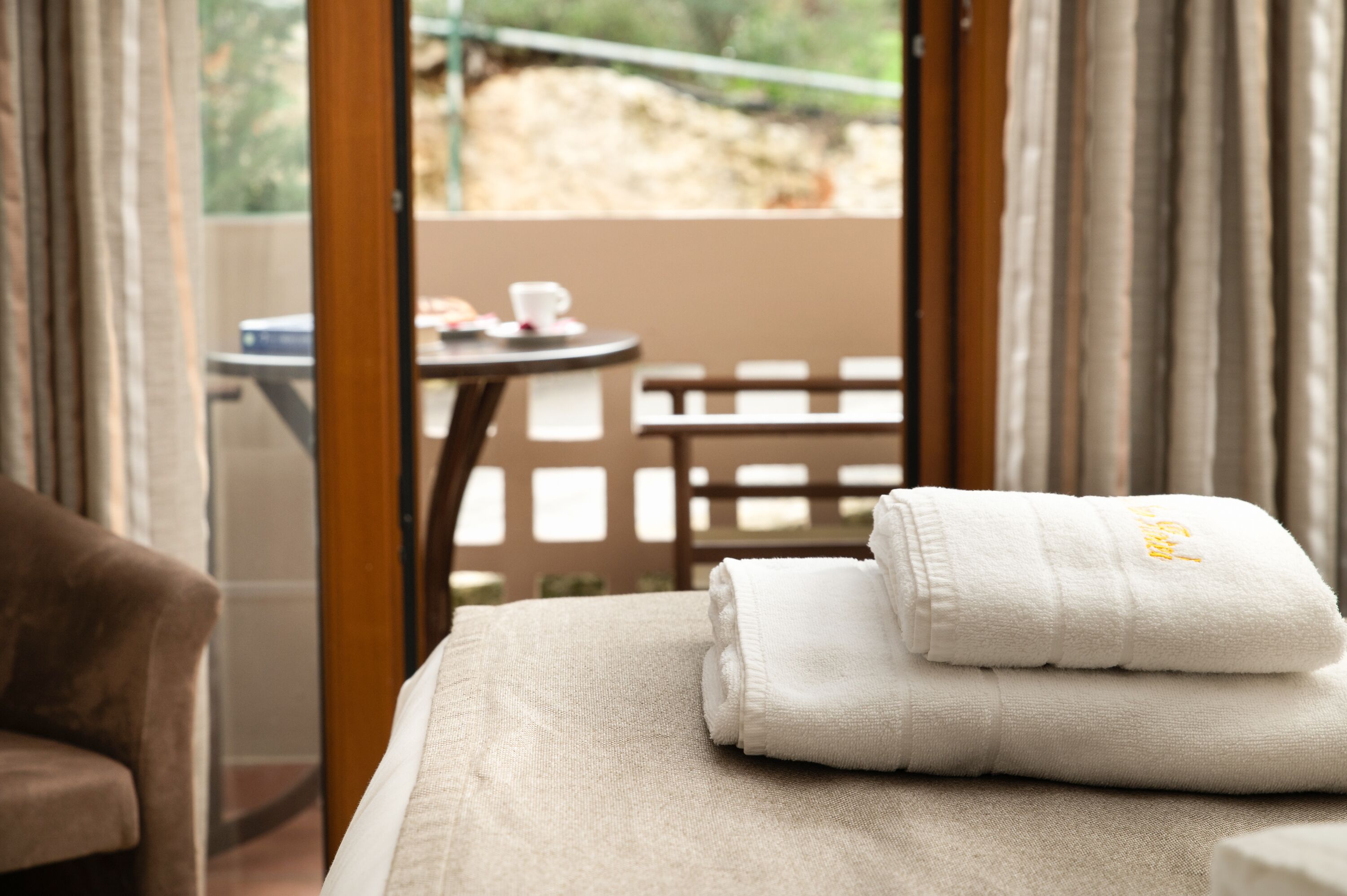 A neatly made bed with folded white towels and a view of an outdoor terrace with a table and chairs.
