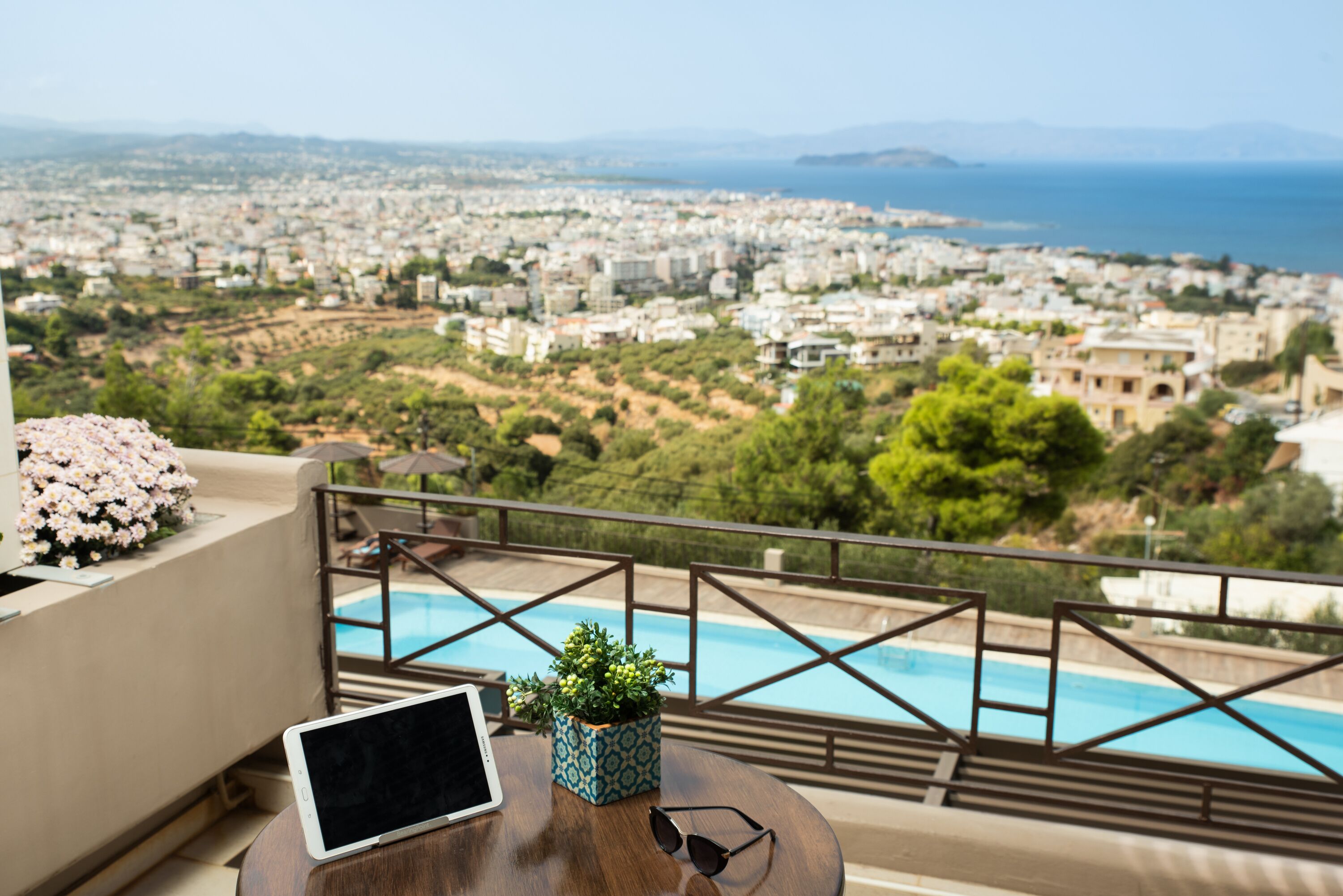 Ein Balkon mit einem Tablet, Sonnenbrille und einem Blumentopf, mit Blick auf eine Stadtlandschaft und einen Pool.