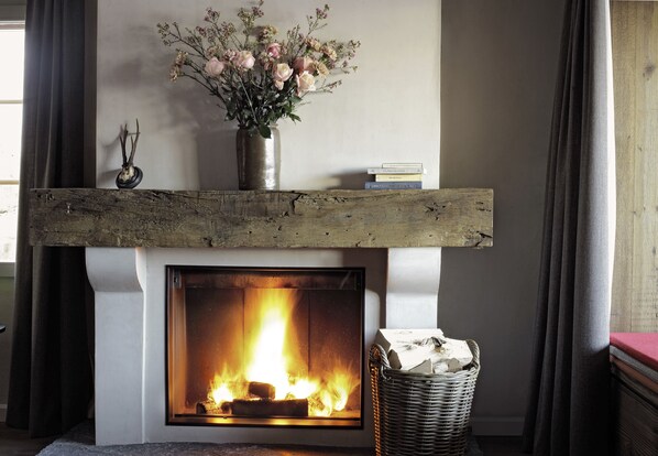 A fireplace with a wooden mantel, a vase with flowers, and a basket of firewood.