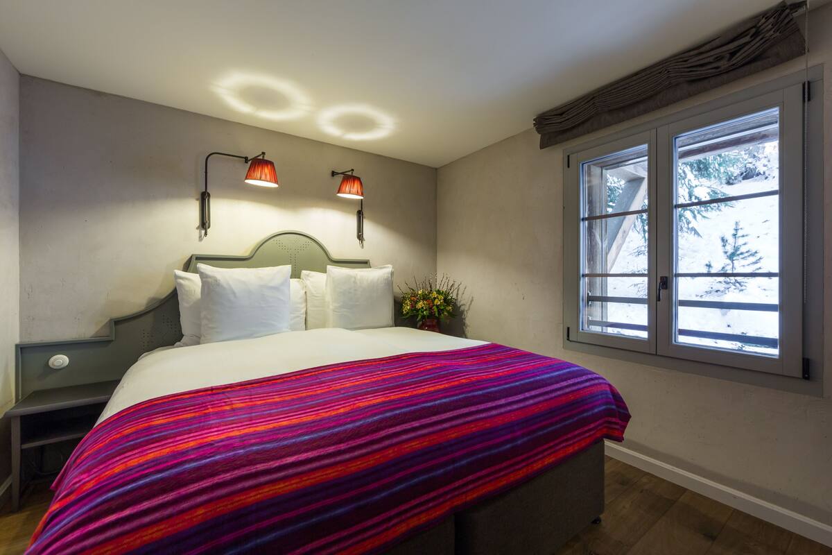 A bedroom with a bed, two bedside lamps, a window with a view of snow, and a decorative wall piece.