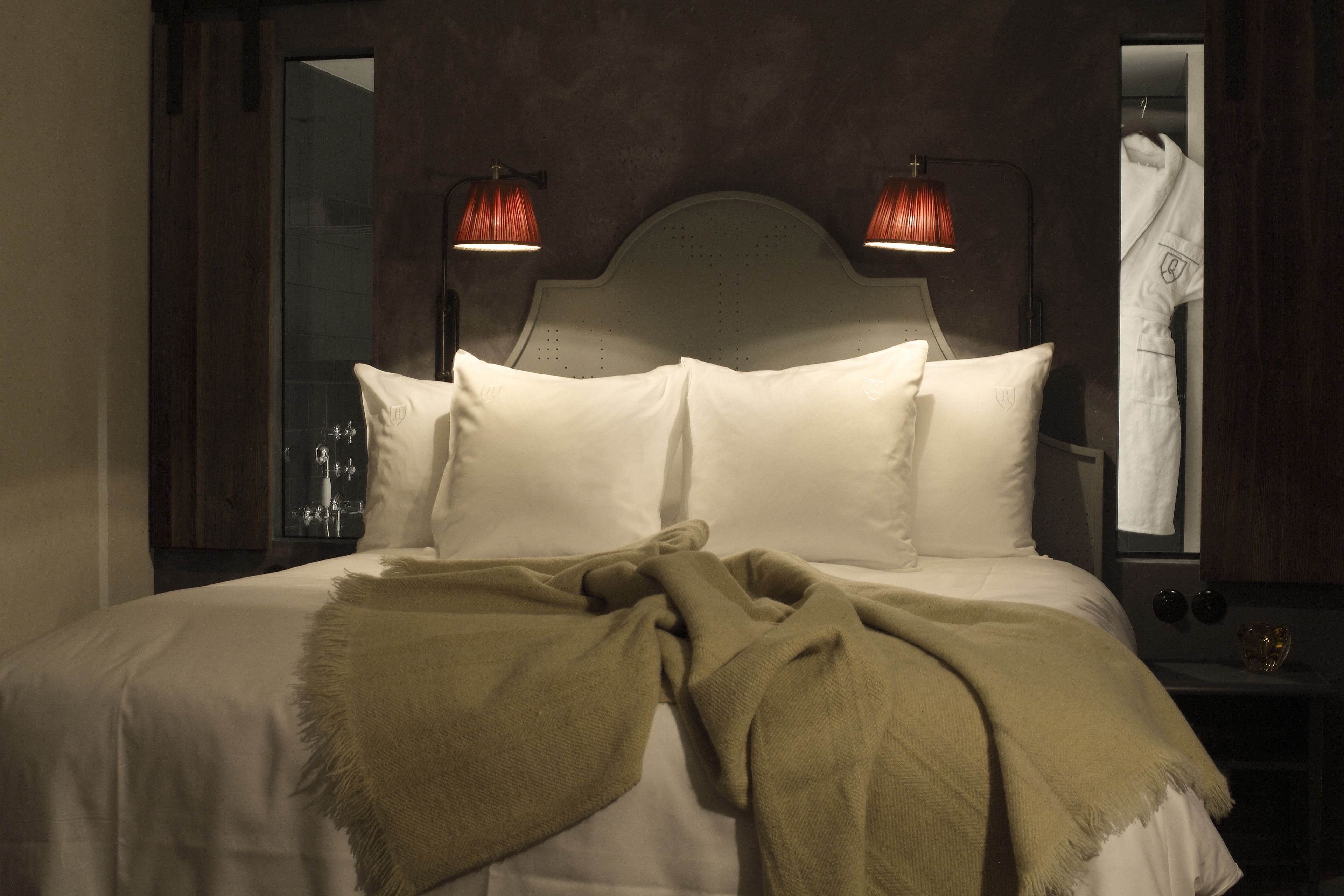 A neatly made bed with white linens and a beige throw blanket, two wall-mounted lamps, and a mirror reflecting a white robe.