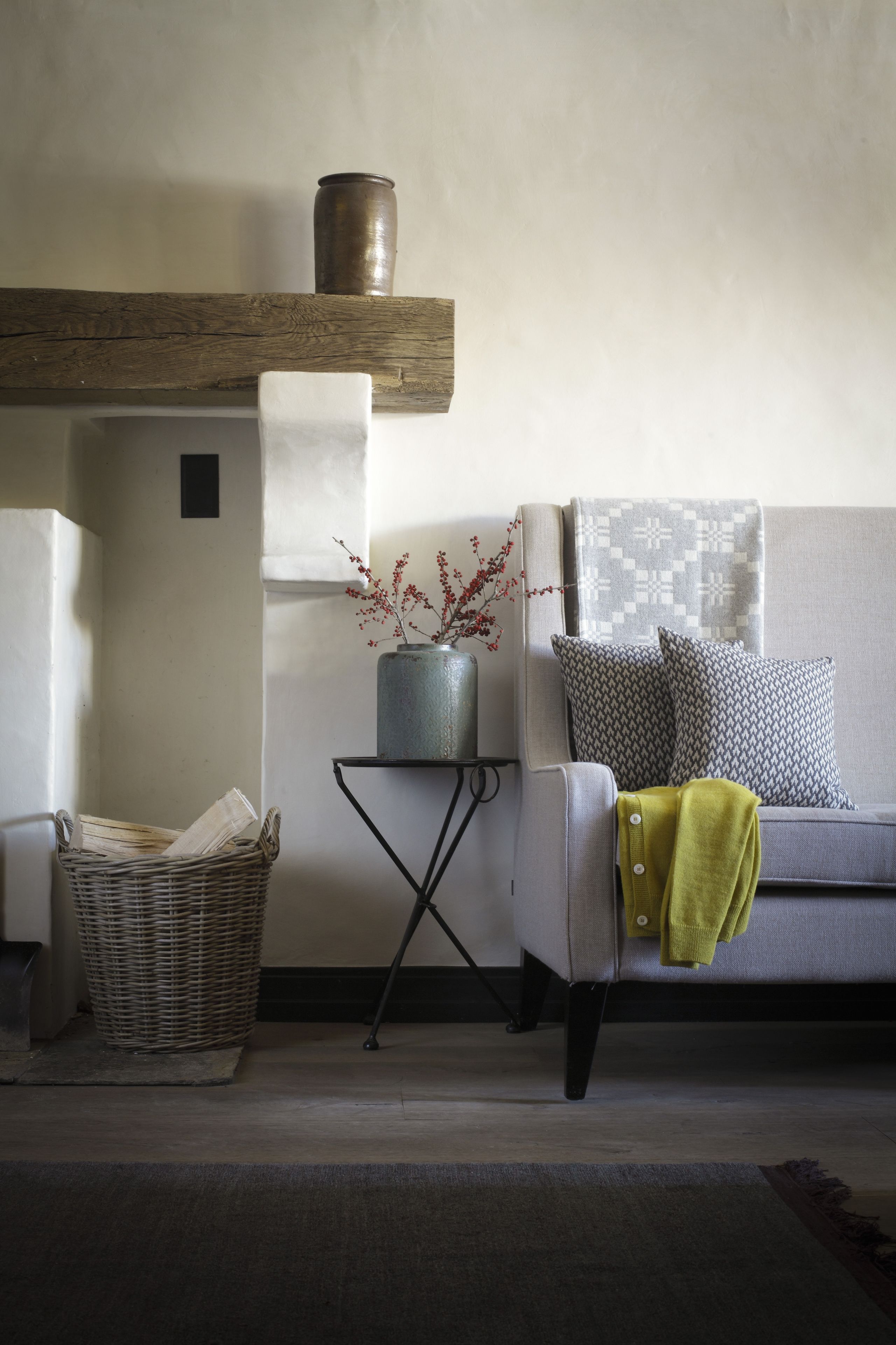 Un salon avec un canapé gris, des coussins à motifs, un plaid vert, un vase avec des fleurs rouges, un panier en osier et une petite table sur laquelle est posé un vase.