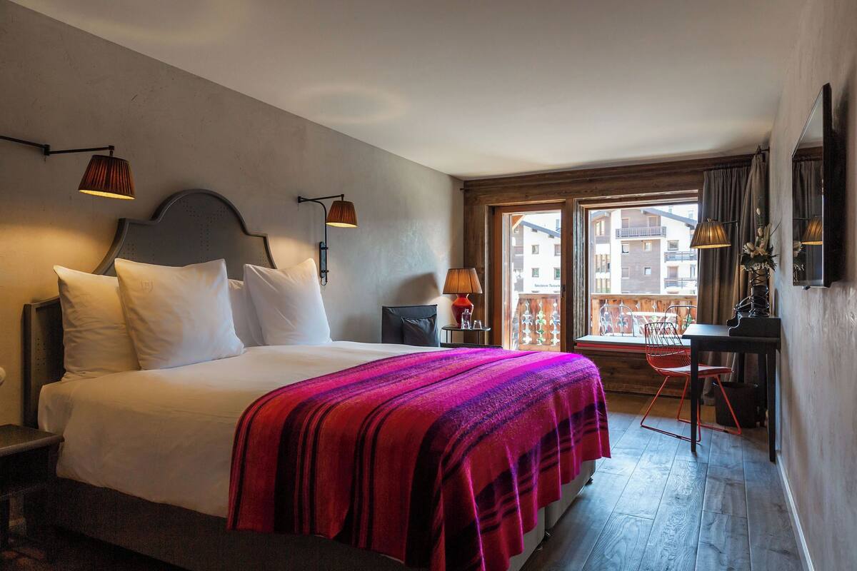 A hotel room with a large bed, a desk with a chair, a window with a view of buildings, and a balcony.