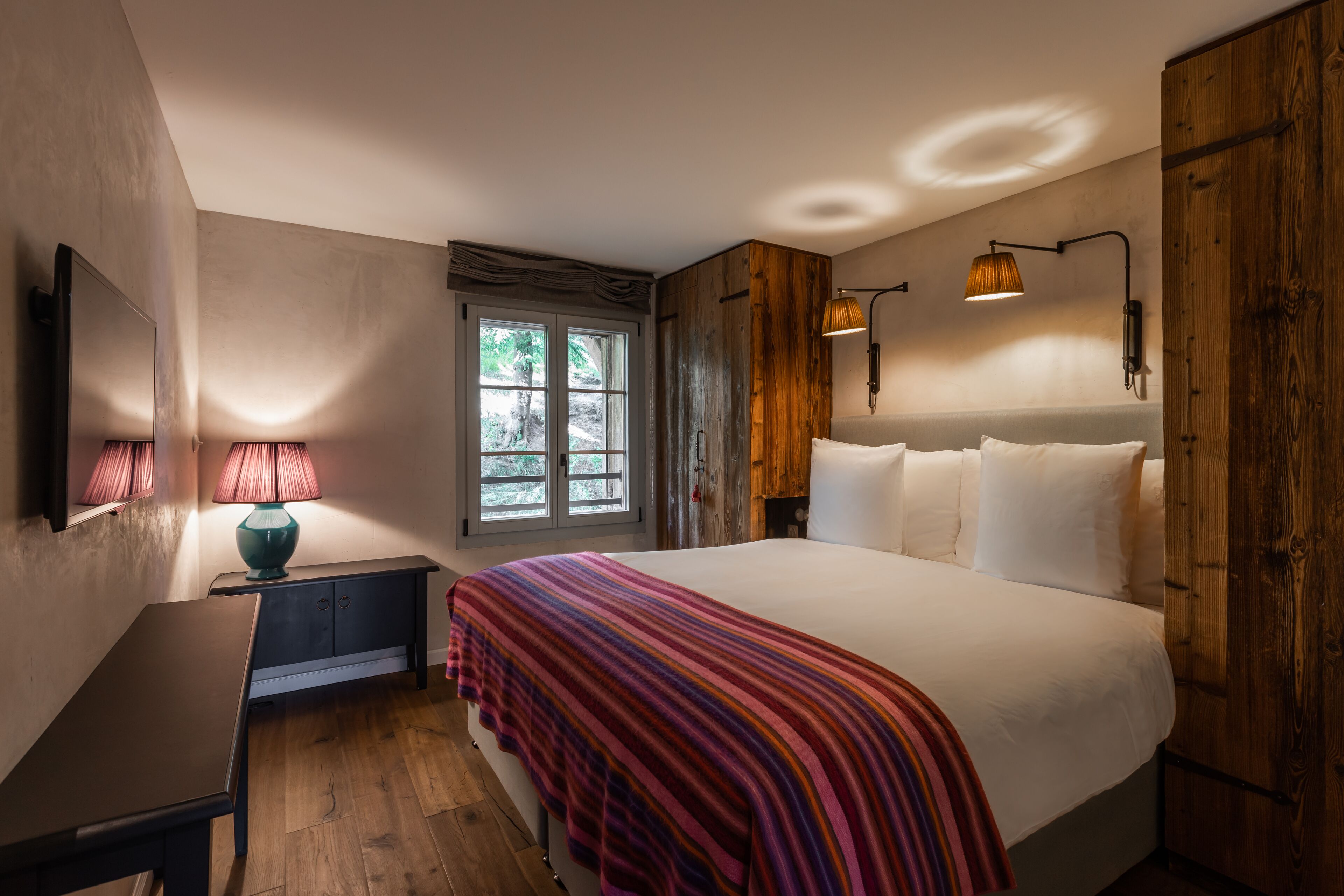 A bedroom with a large bed, a desk, a window with curtains, and a wooden wardrobe.