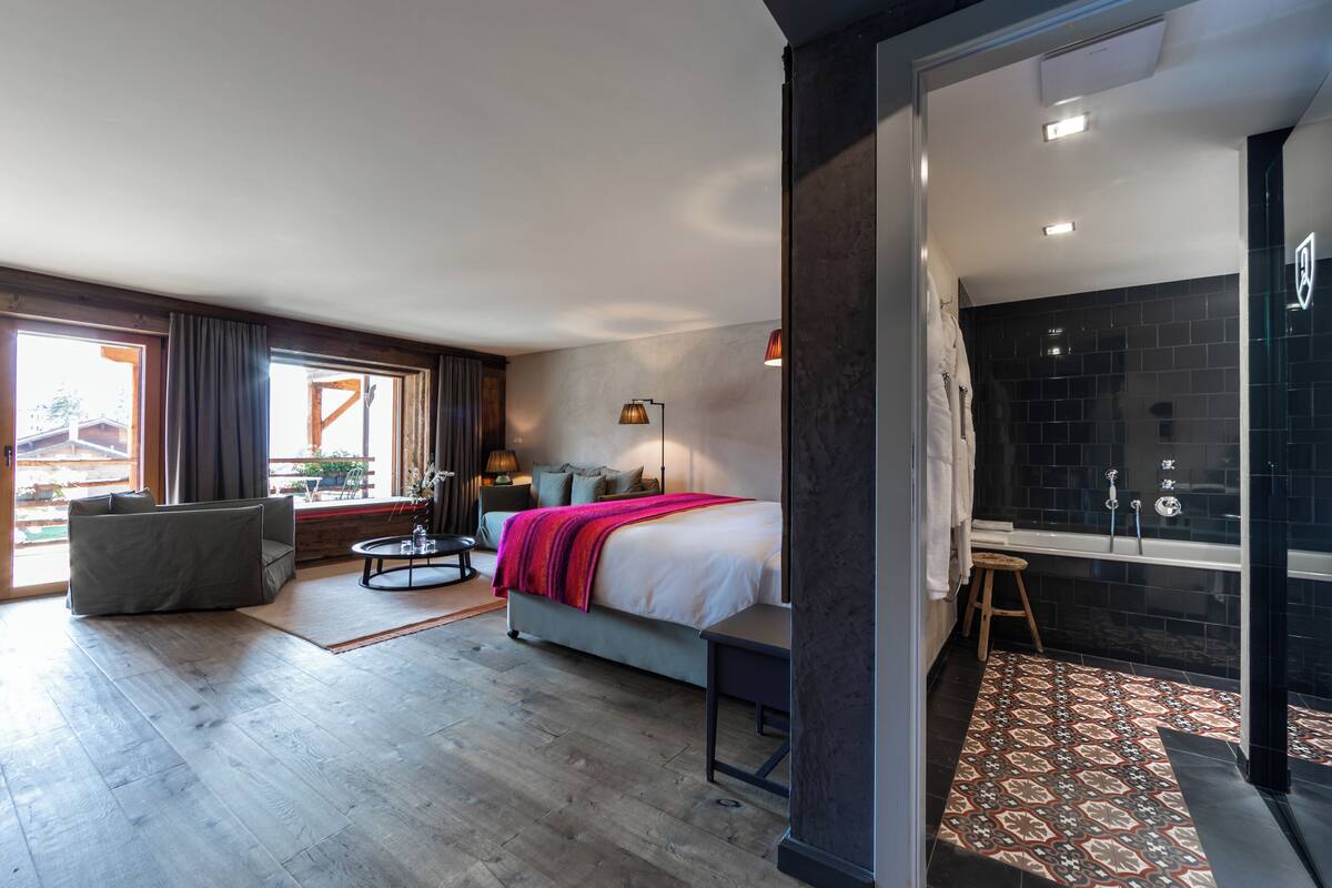 A modern hotel room with a large bed, a sitting area with a sofa and a coffee table, a small round table with a lamp, and a bathroom with a glass shower enclosure.