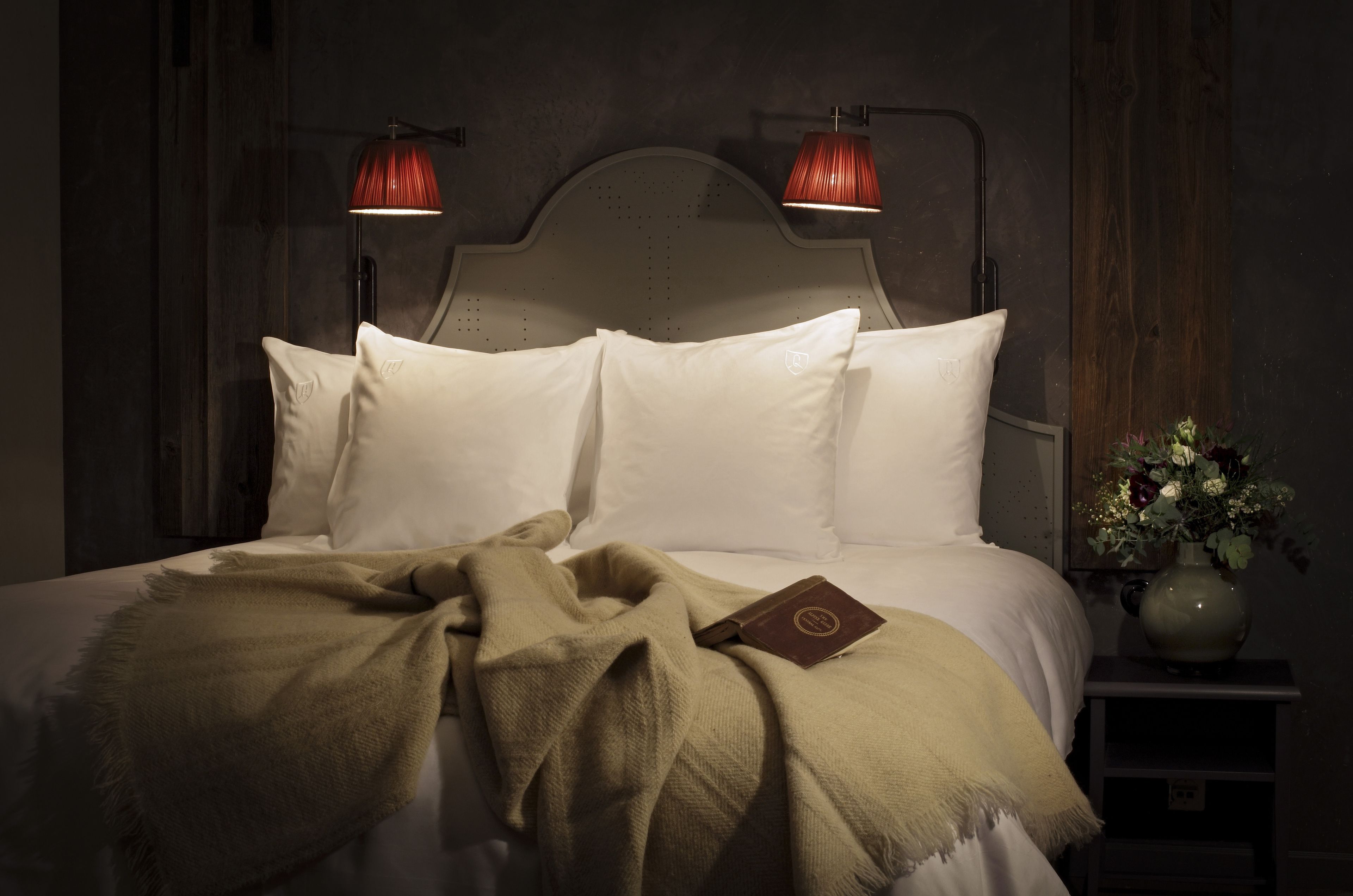 A neatly made bed with white linens and a beige throw blanket, two bedside lamps, and a vase with flowers on a nightstand.
