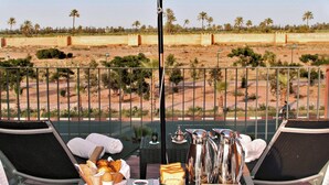 Balcón con mesa dispuesta para el desayuno, que incluye pan, huevos y bebidas, con vistas a un paisaje desértico con palmeras.