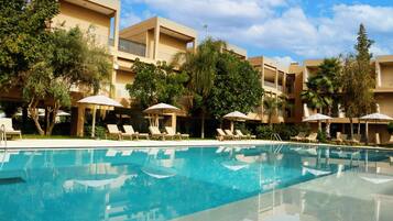 Una piscina con sillas de descanso y sombrillas, rodeada de árboles y un edificio con balcones.