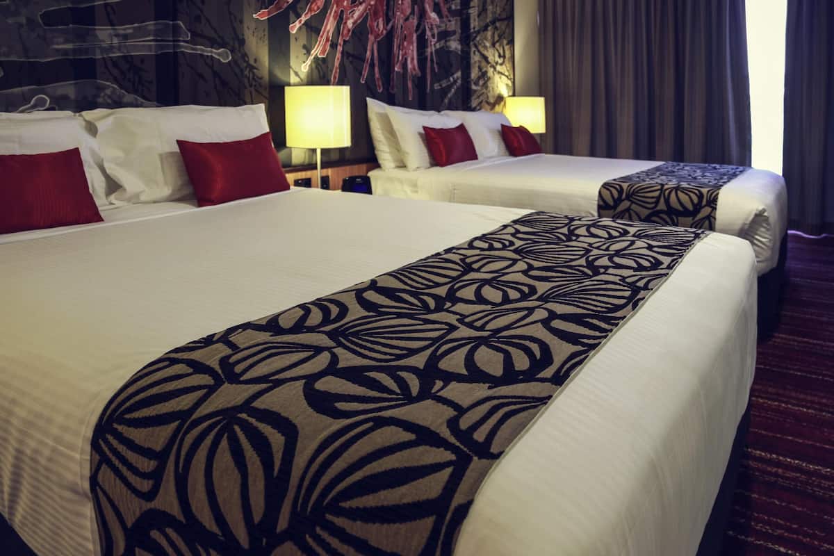 Une chambre d’hôtel avec deux lits, chacun agrémenté de draps blancs et d’oreillers rouges, d’un plaid à motifs, d’un mur sombre orné d’œuvres d’art abstraites et d’une lampe sur une table de chevet.