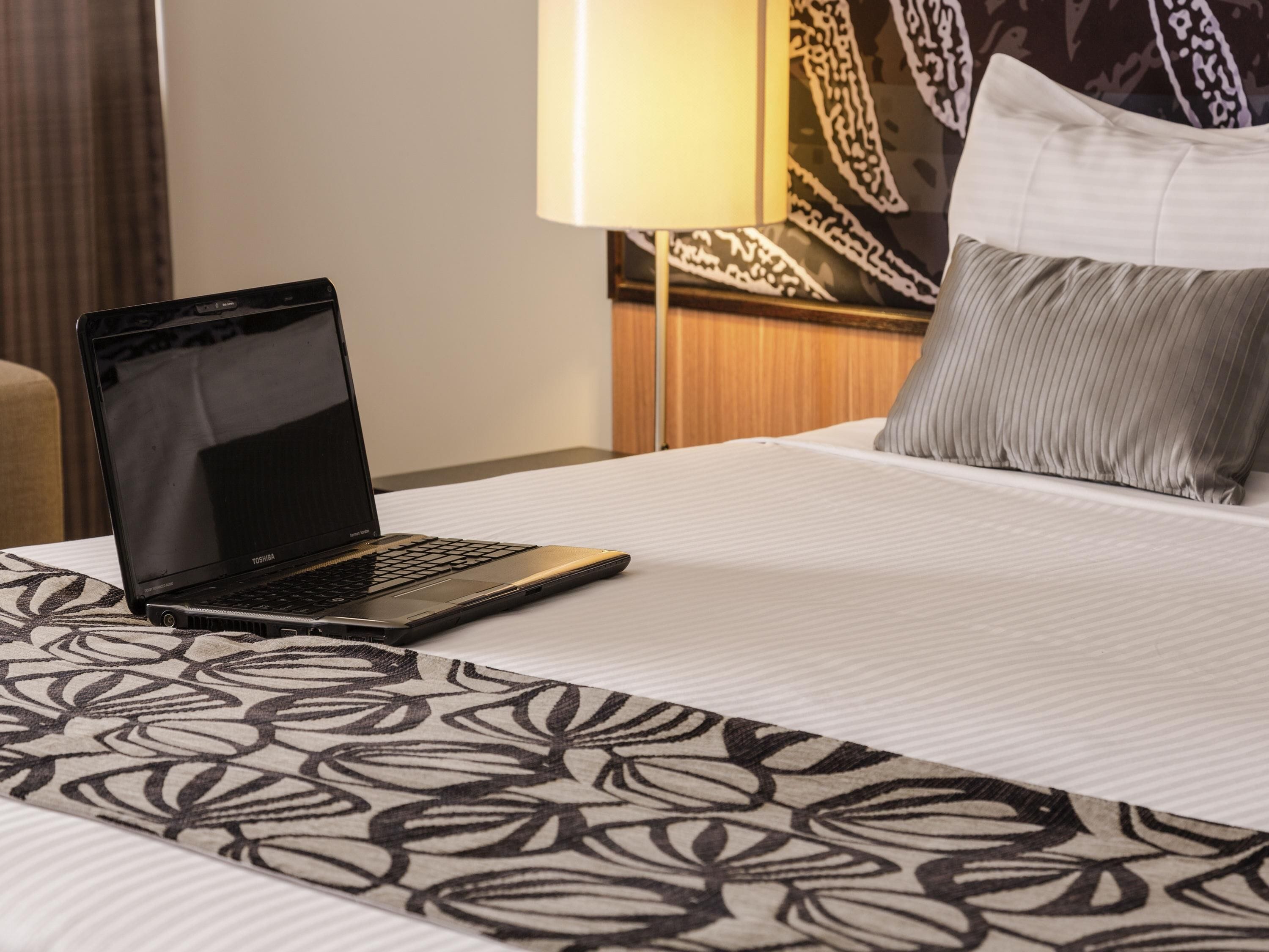Une chambre d’hôtel avec un lit, un ordinateur portable posé sur le lit, une couvre-lit à motifs, un oreiller, une lampe et un mur orné d’un motif décoratif.