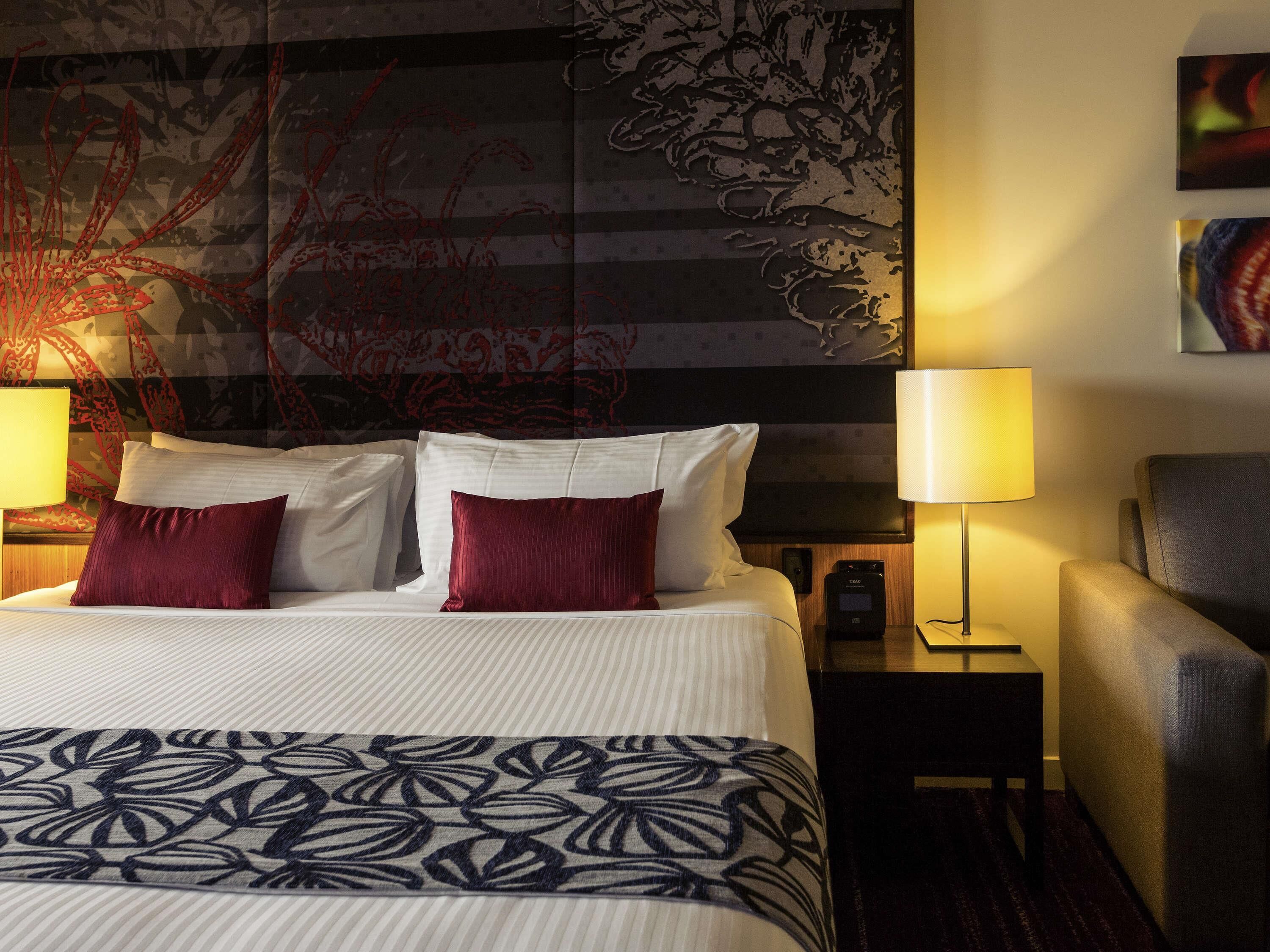 A hotel room with a large bed, two bedside lamps, a chair, a nightstand with a phone, and a wall with a large abstract painting.