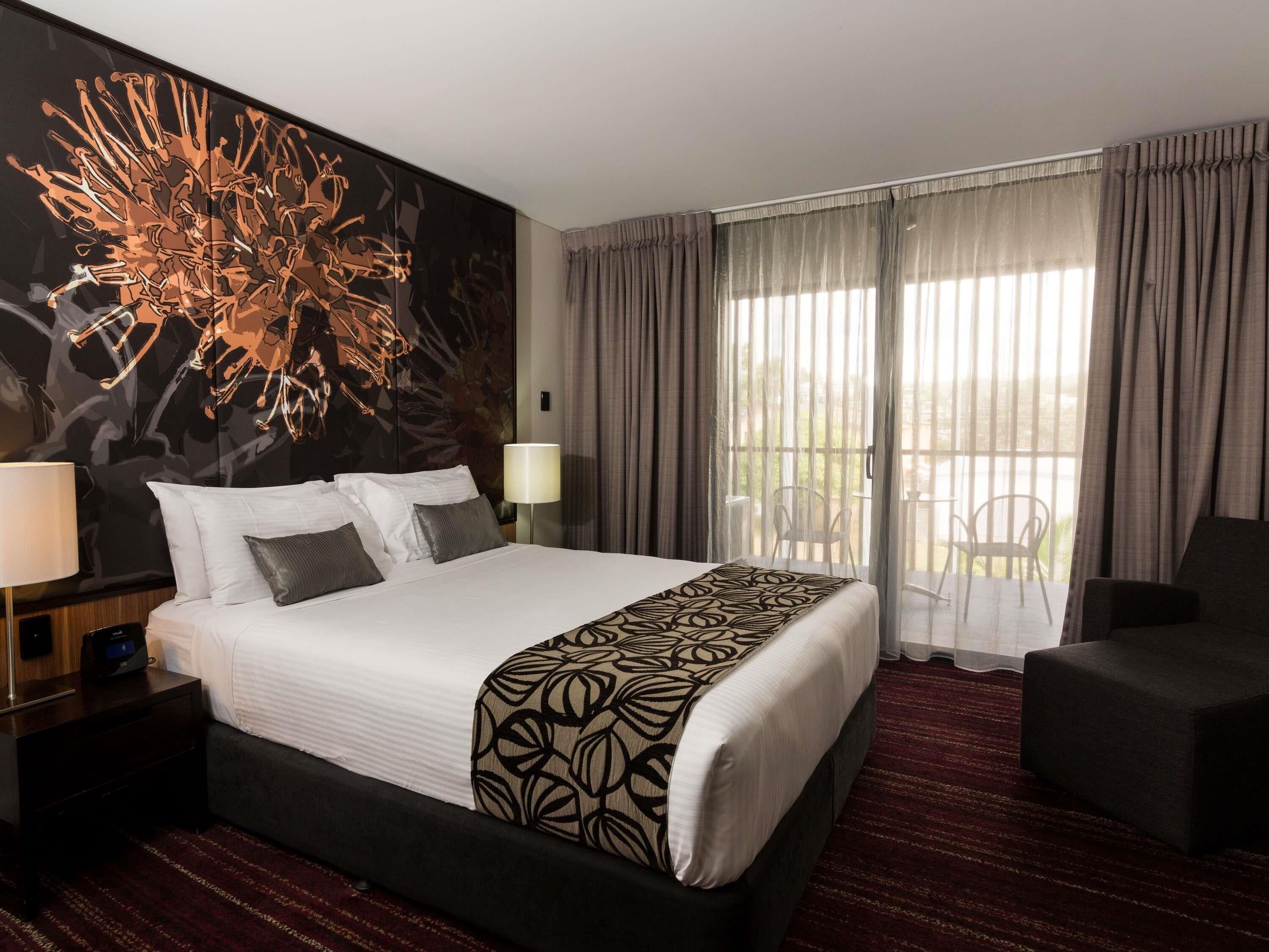 A hotel room with a large bed, a nightstand, a lamp, a chair, and a balcony with a view.