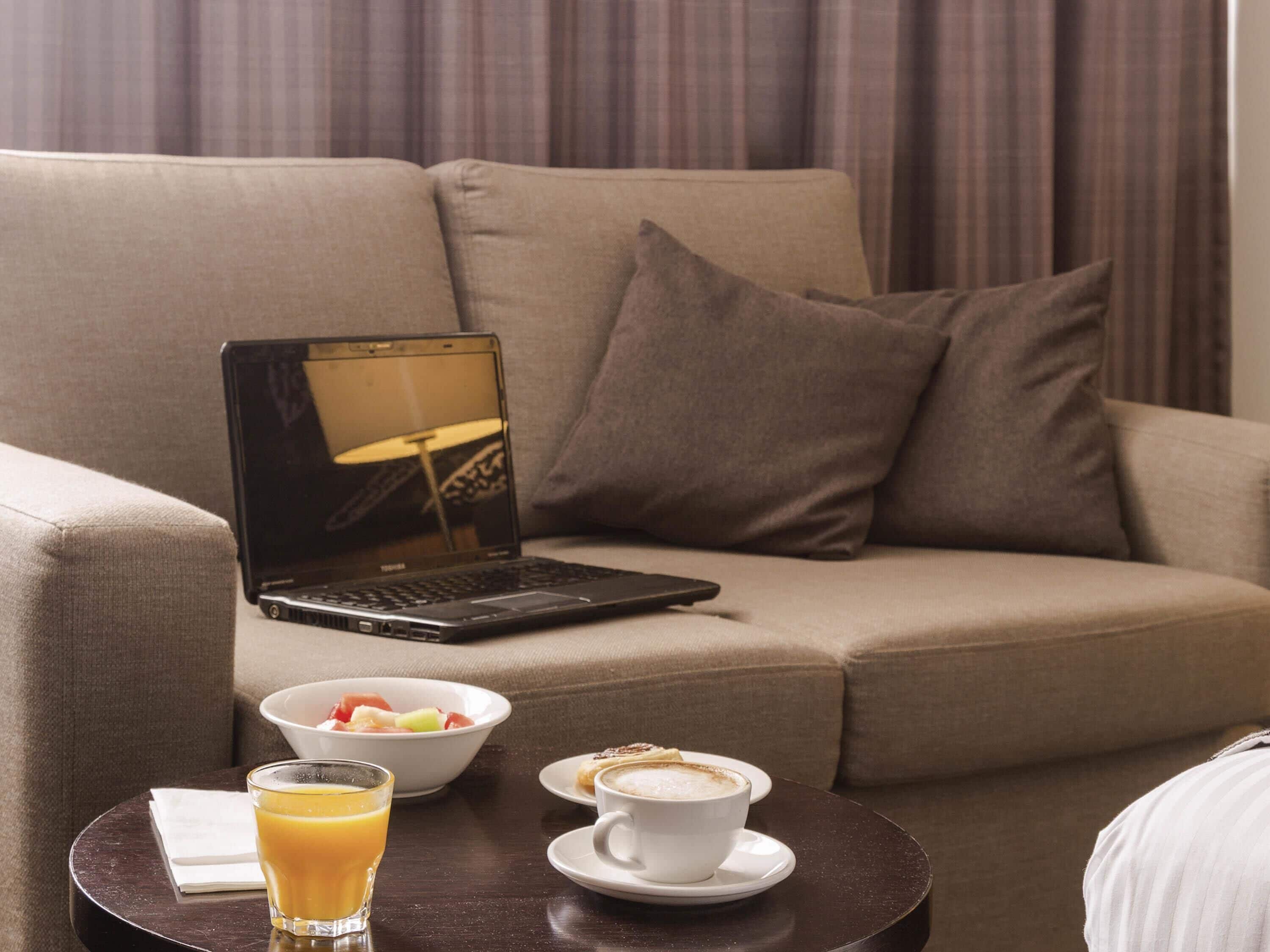 Un ordinateur portable sur un canapé, un verre de jus d’orange, un saladier de fruits et une tasse de café sur une petite table.
