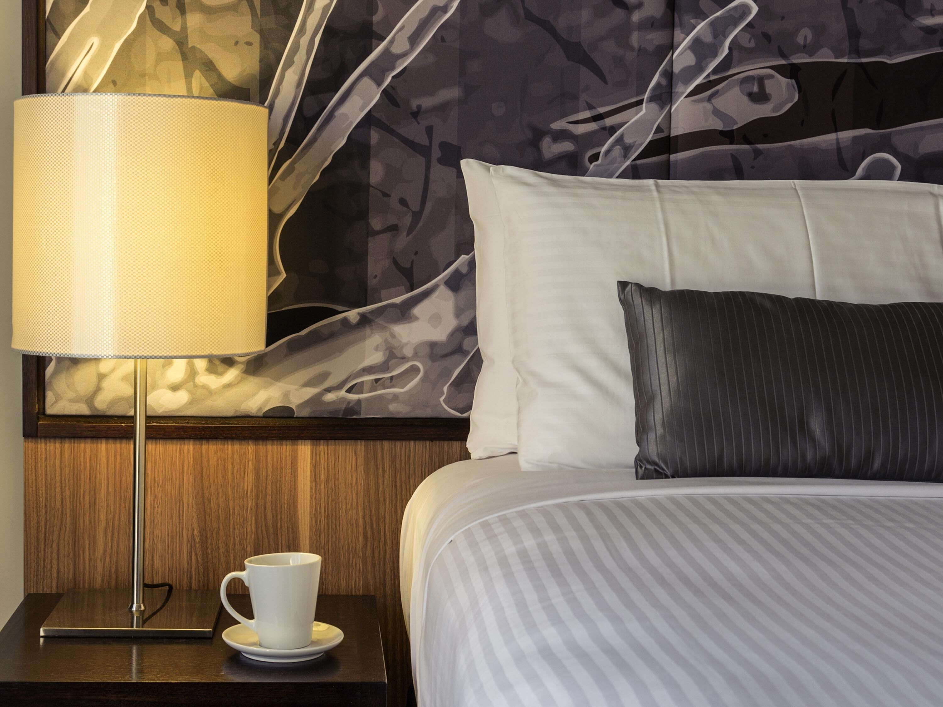 A neatly made bed with a striped comforter, a dark pillow, a white pillow, a lamp with a beige shade, and a white mug on a wooden nightstand.