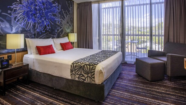 Habitación de hotel con una cama grande, dos lámparas de noche, una mesita de noche, una silla, un balcón con mesa y un cuadro abstracto de gran tamaño en la pared.