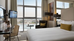 A hotel room with a large bed, a desk with a computer, a chair, a TV, and a view of a cityscape through large windows. - Centro Capital Centre (Abu Dhabi)