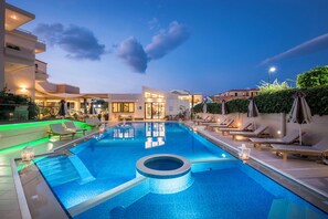A swimming pool with lounge chairs and a circular hot tub, surrounded by modern buildings and greenery. - Oscar Suites & Village (Chania)
