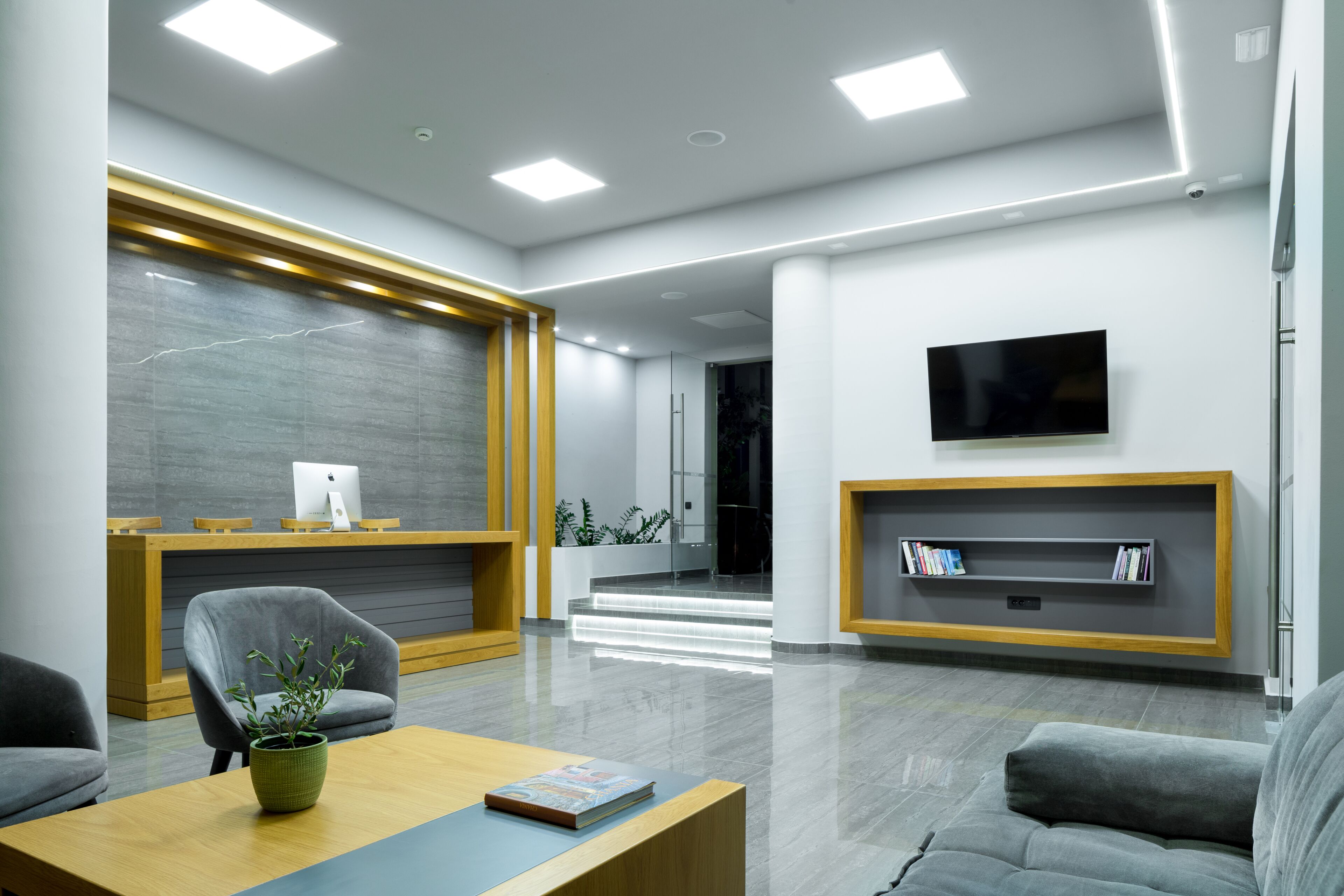 A modern reception area with a wooden reception desk, a grey armchair, a wooden coffee table with a book, a flat-screen TV mounted on the wall, and a bookshelf with books.