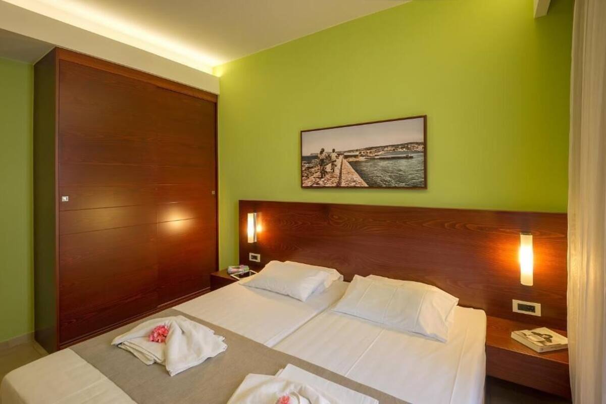 A hotel room with two beds, a wooden wardrobe, a nightstand with a lamp, and a framed picture on the wall.