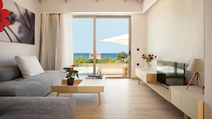 A modern living room with a sofa, coffee table, TV, and a view of the sea through large sliding doors. - Oscar Suites & Village (Chania)