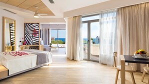 A modern hotel room with a bed, a dining table with chairs, a balcony with a view of the sea, and a ceiling fan. - Oscar Suites & Village (Chania)