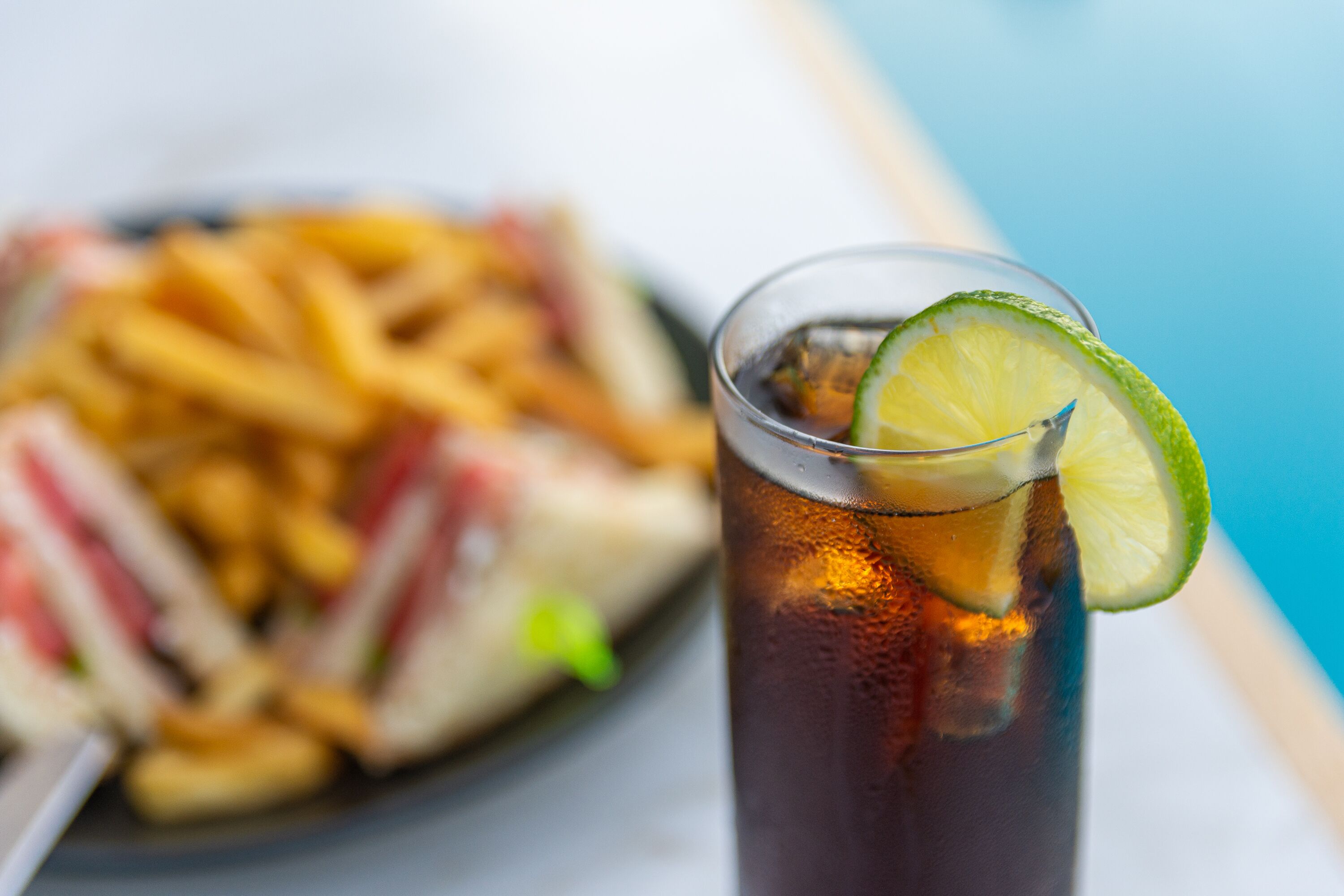 A glass of iced tea with a lime slice, next to a plate of French fries and a sandwich.