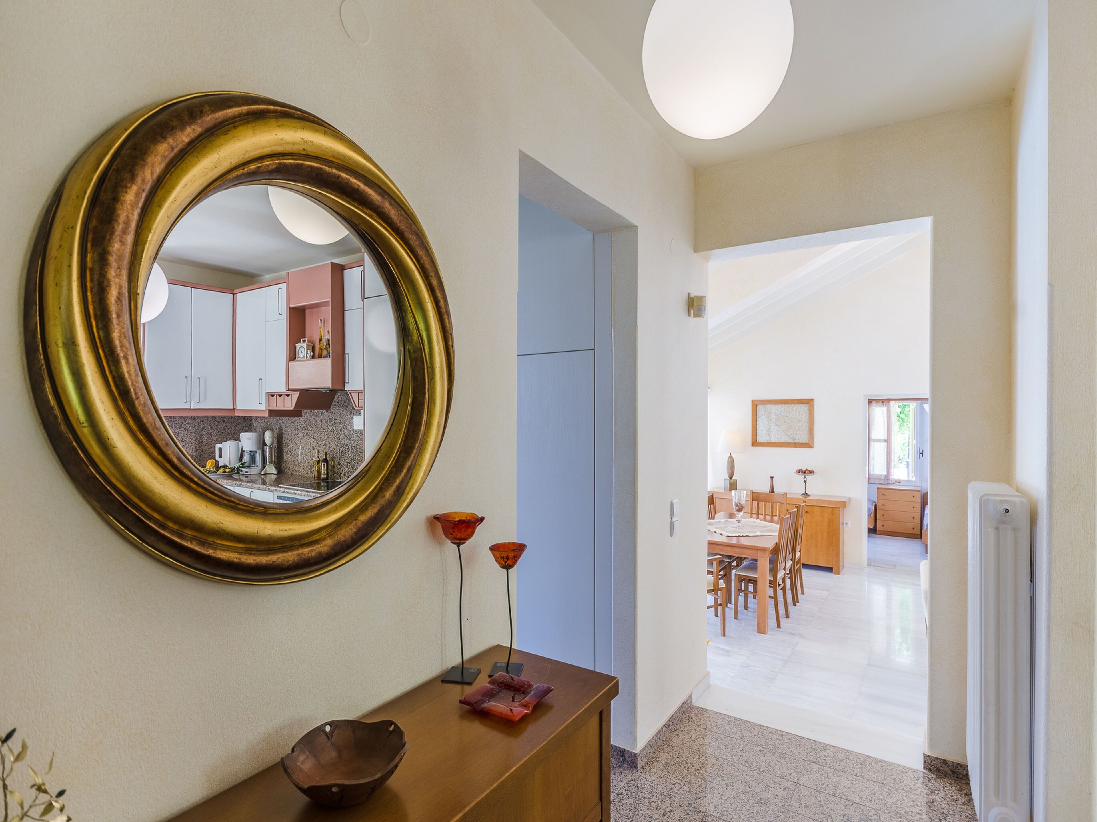 Un pasillo con un espejo grande, una consola de madera con un cuenco y un jarrón rojo, y vista hacia un comedor con una mesa y sillas de madera.