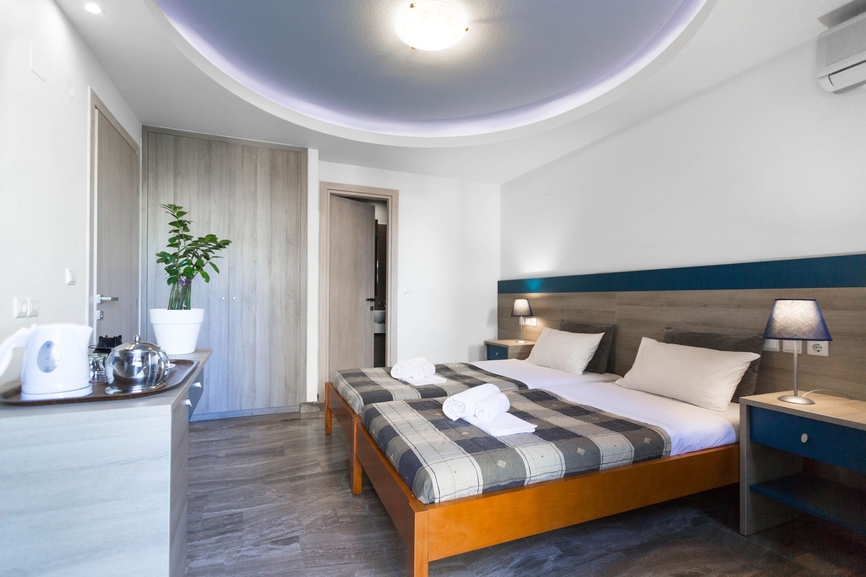 A modern hotel room with a wooden bed, a bedside table with a lamp, a potted plant, and a small kitchenette area.