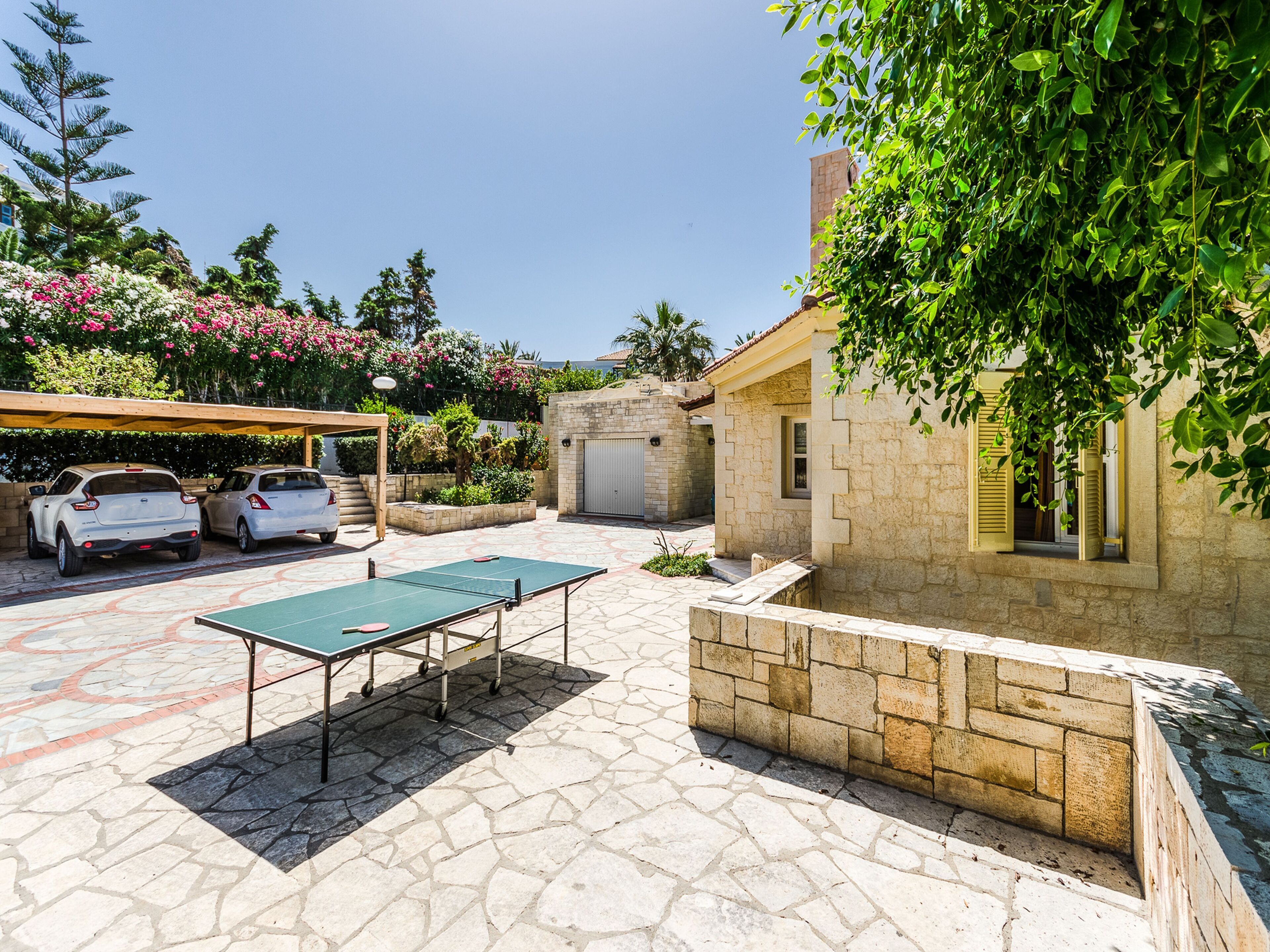Un patio con mesa de ping pong, dos autos y un edificio de piedra con ventana y contraventanas.