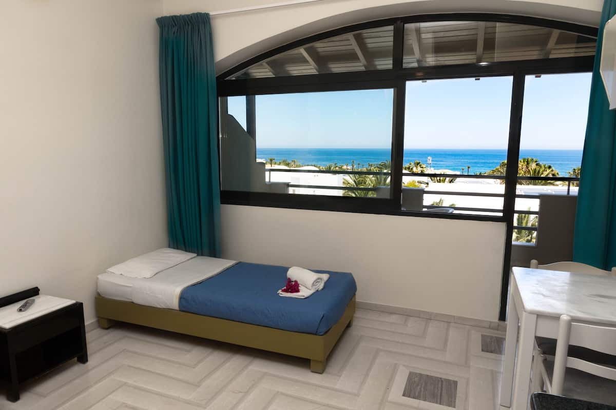 A room with a bed, a small table, and a window with a view of the ocean.