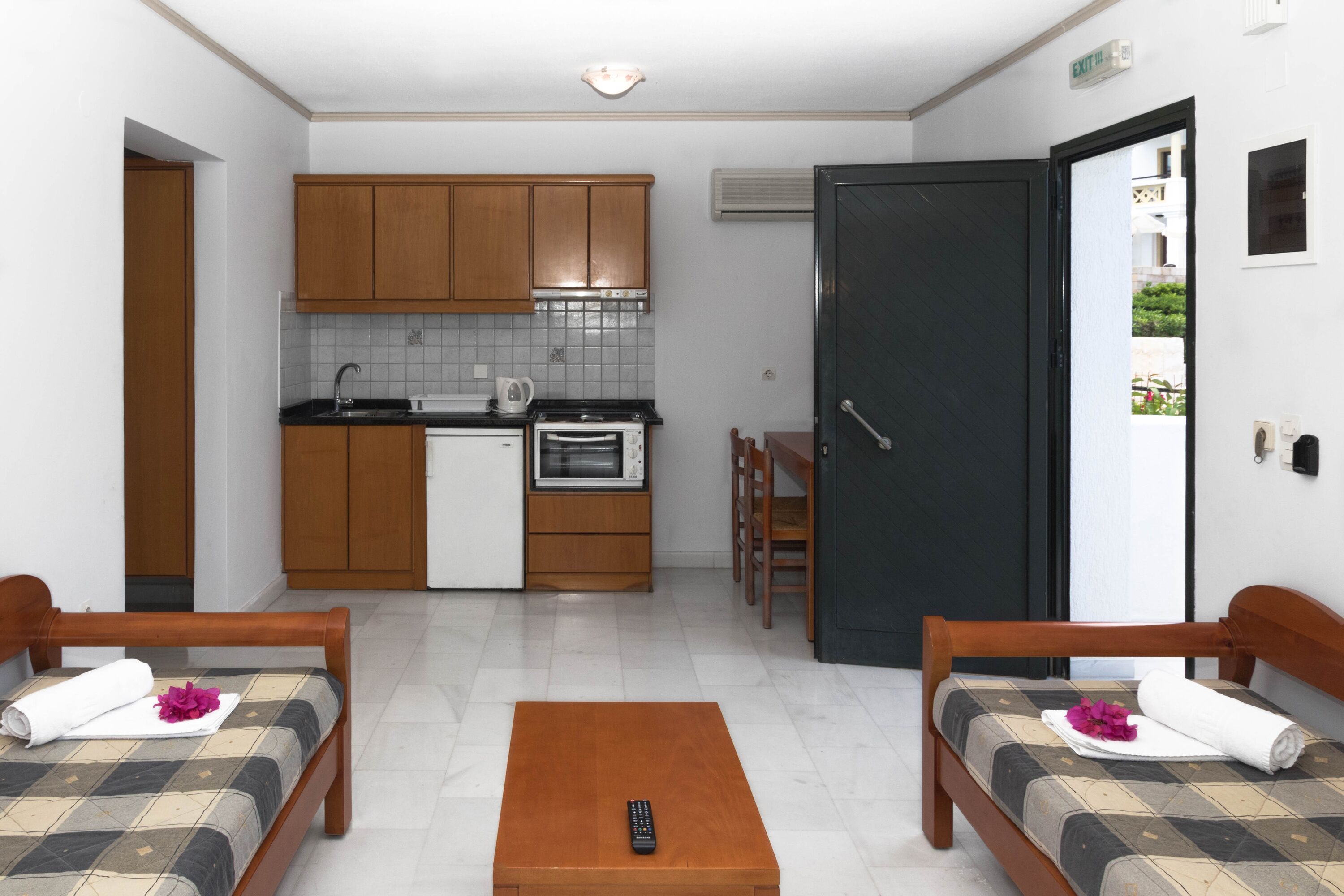 Une chambre d’hôtel compacte comprenant un coin cuisine, deux lits simples, une petite table en bois et une porte donnant sur l’extérieur.