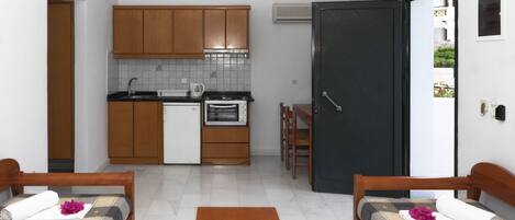 A compact hotel room with a kitchenette, two single beds, a small wooden table, and a door leading to the outside.